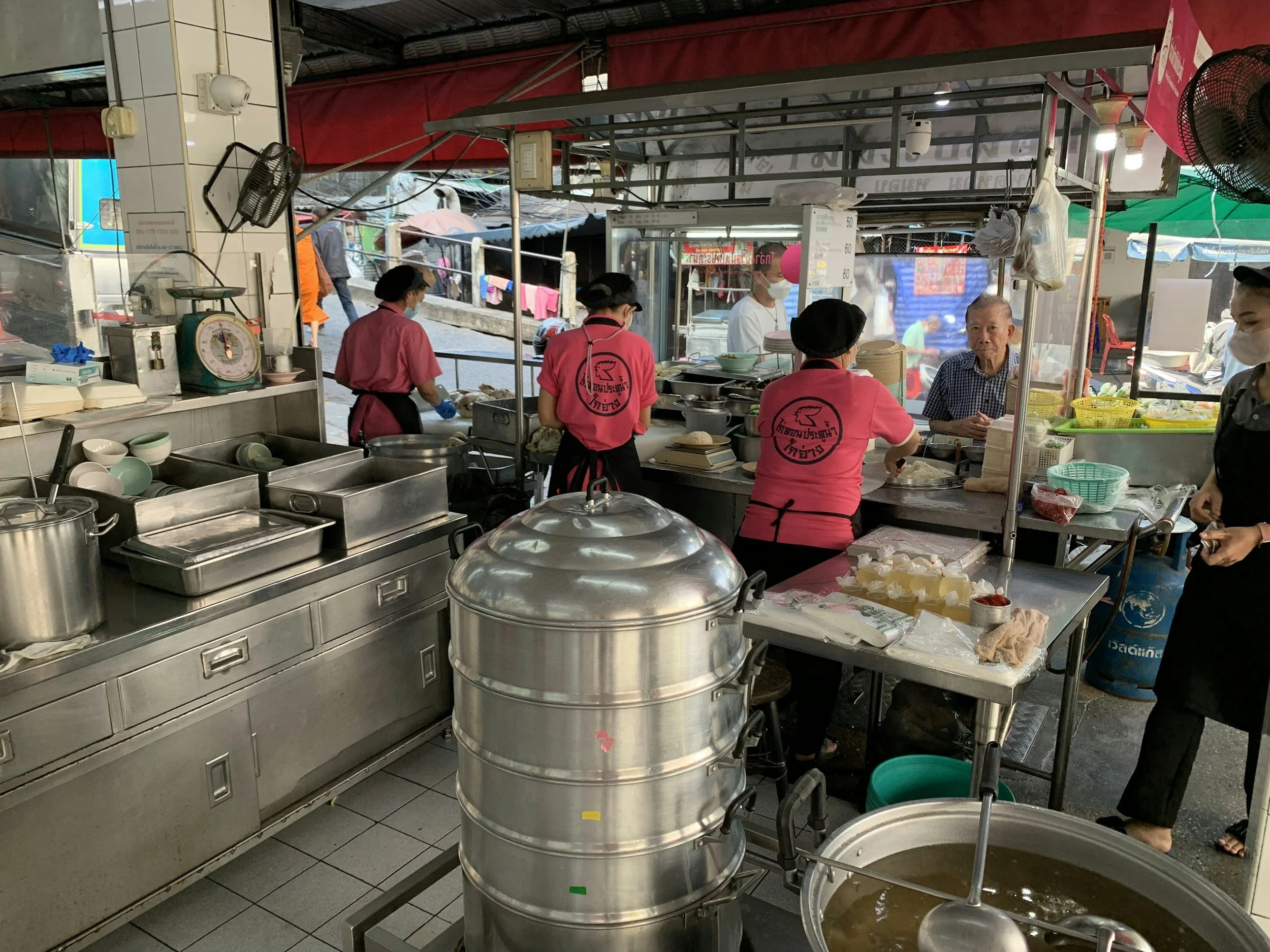 A busy street food stall with four staff wearing pink shirts and black aprons, preparing food for customers. Several bowls, steamers, and cooking utensils are visible, along with a scale and a fan. An elderly man is talking to one of the staff, and another person is seen behind the counter. The stall is outdoors with colorful tents and passersby in the background.