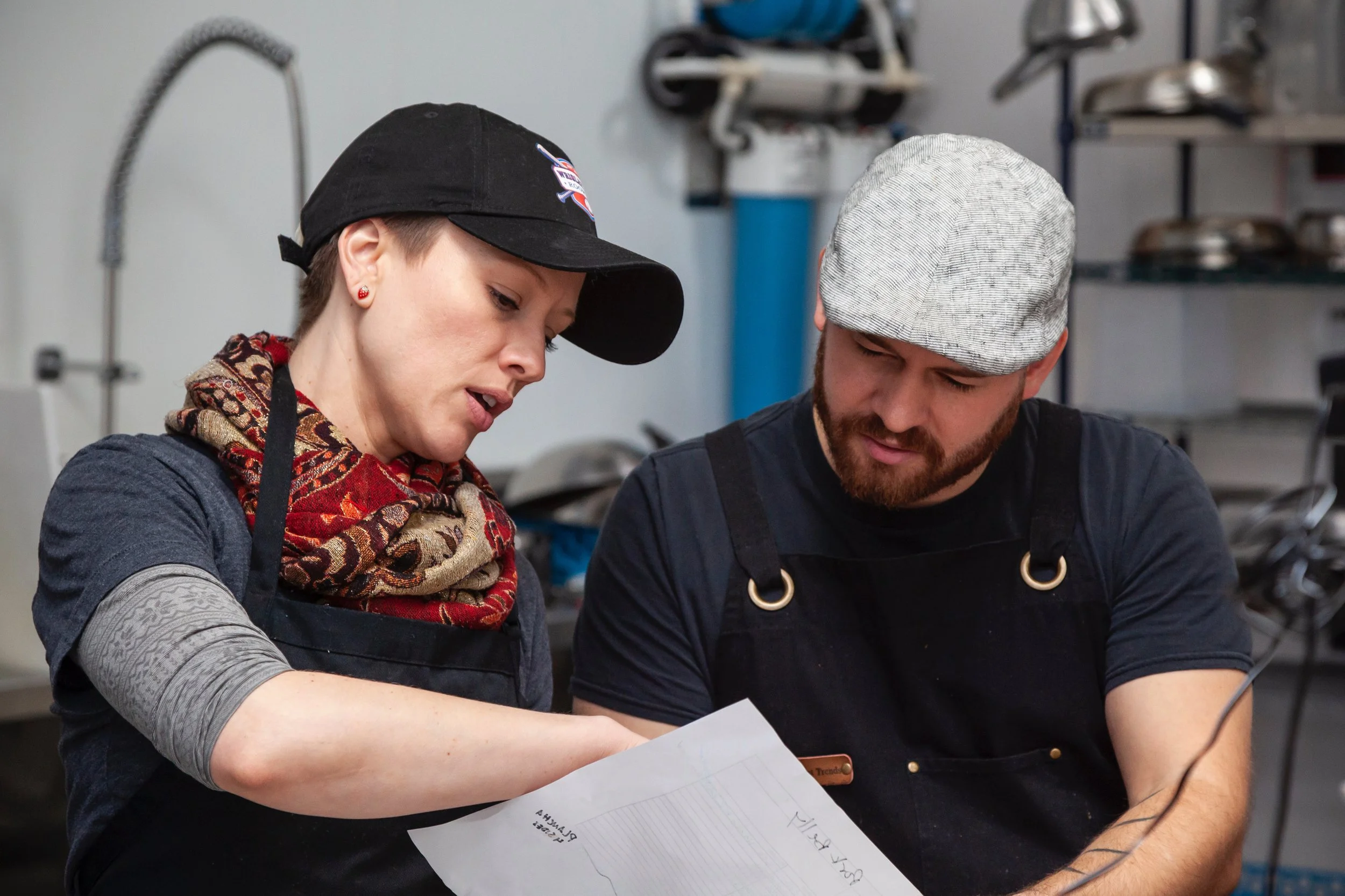 A woman and man in a workshop reviewing a paper together. The woman is wearing a black baseball cap, patterned scarf, and apron, and the man is wearing a flat cap and apron. Shelves and tools are in the background.
