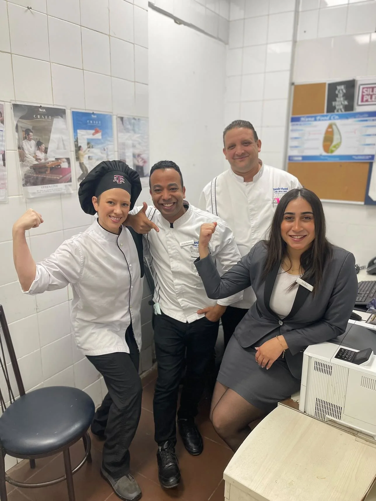 Four restaurant staff members, three men and one woman, posing together in a kitchen. Two of them are flexing their arms. They are all smiling and dressed in uniforms.
