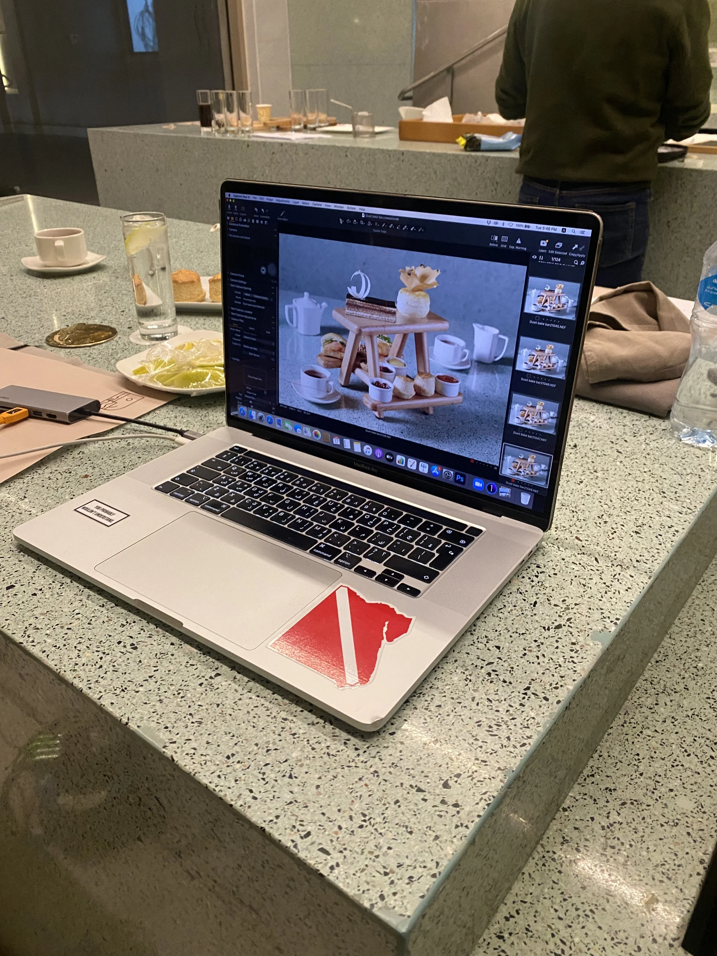 A laptop on a kitchen or dining table with a photo editing software open, displaying a photo of a wooden tray with tea and snack items. Surrounding the laptop are a glass of water, a cup with a saucer, a plate with lemon slices, and some snacks. There are also some papers, a water bottle, and a person standing in the background wearing a green jacket.