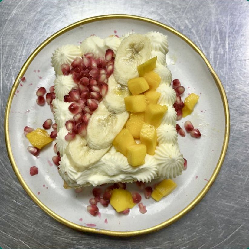 A slice of cake with whipped cream, topped with pomegranate seeds, banana slices, and mango chunks, on a white plate with a gold rim.
