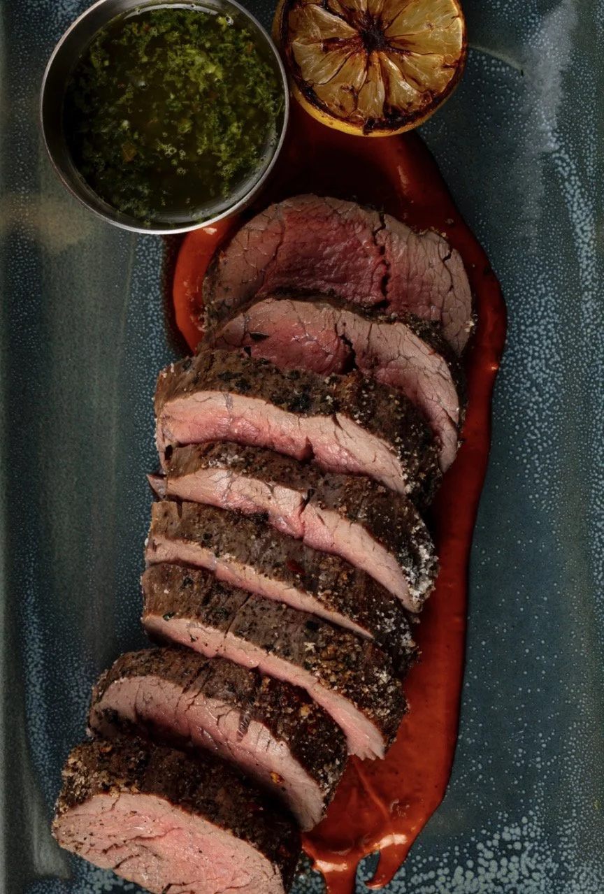 Sliced roasted beef tenderloin on a platter with a side of chimichurri sauce, a halved lemon, and a small bowl of green sauce.