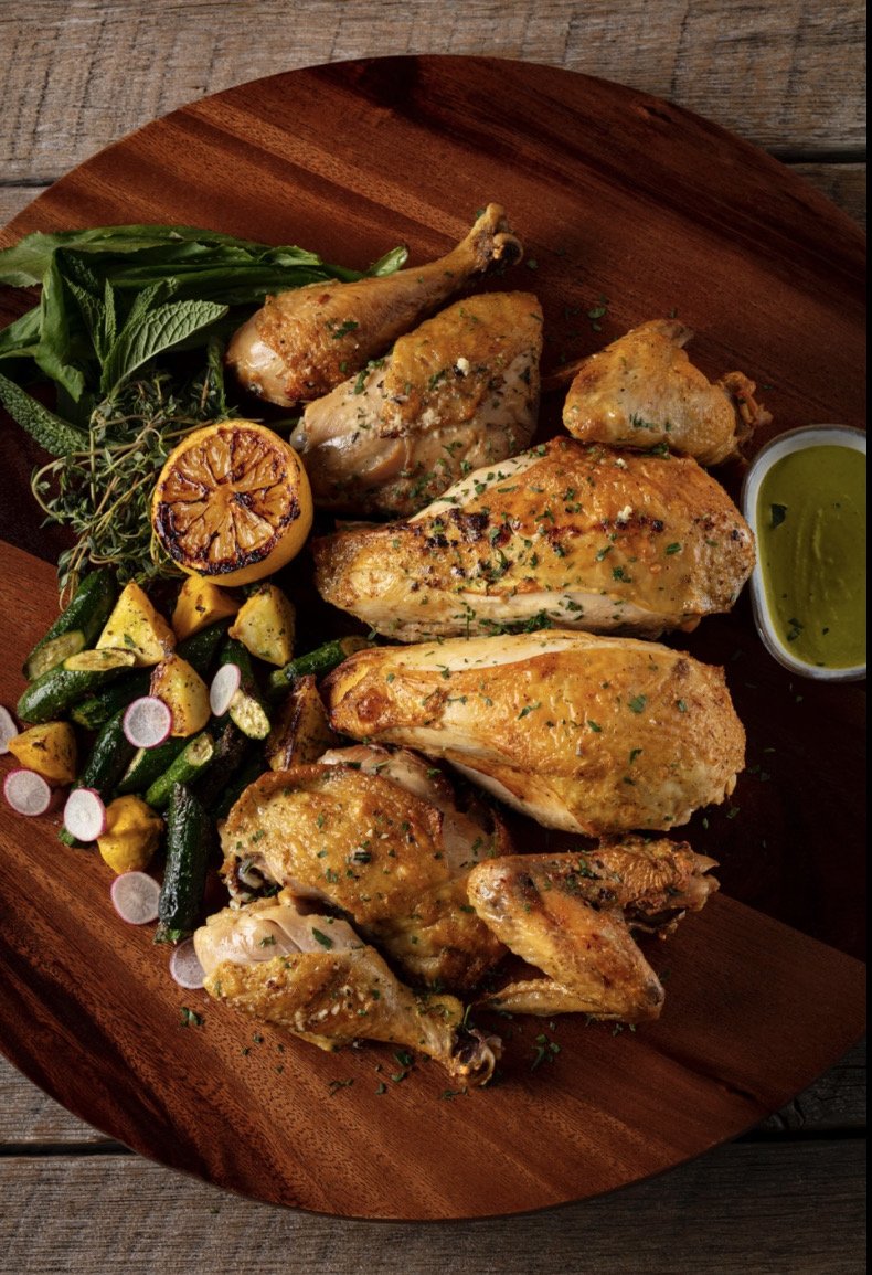 A wooden platter with roasted chicken pieces, grilled vegetables, lemon half, herbs, and green sauce.