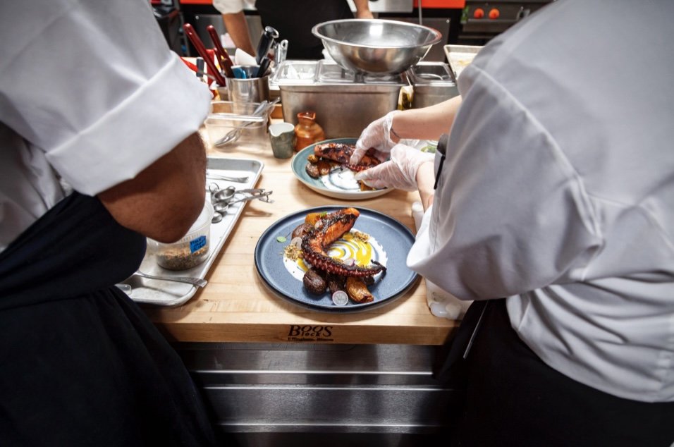Chefs preparing grilled octopus on a blue plate in a professional kitchen.