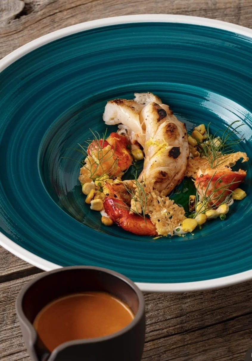 A plated gourmet seafood dish with lobster and fish, garnished with herbs and vegetables, served on a teal ceramic plate with a wooden table underneath.