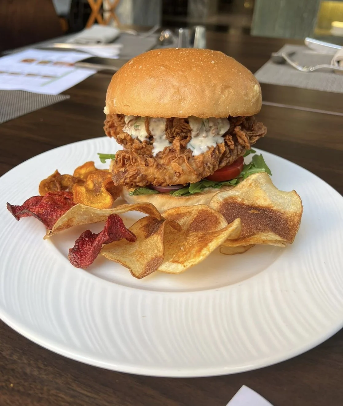 Fried chicken sandwich with lettuce, tomato, and sauce, served with potato chips on a white plate.