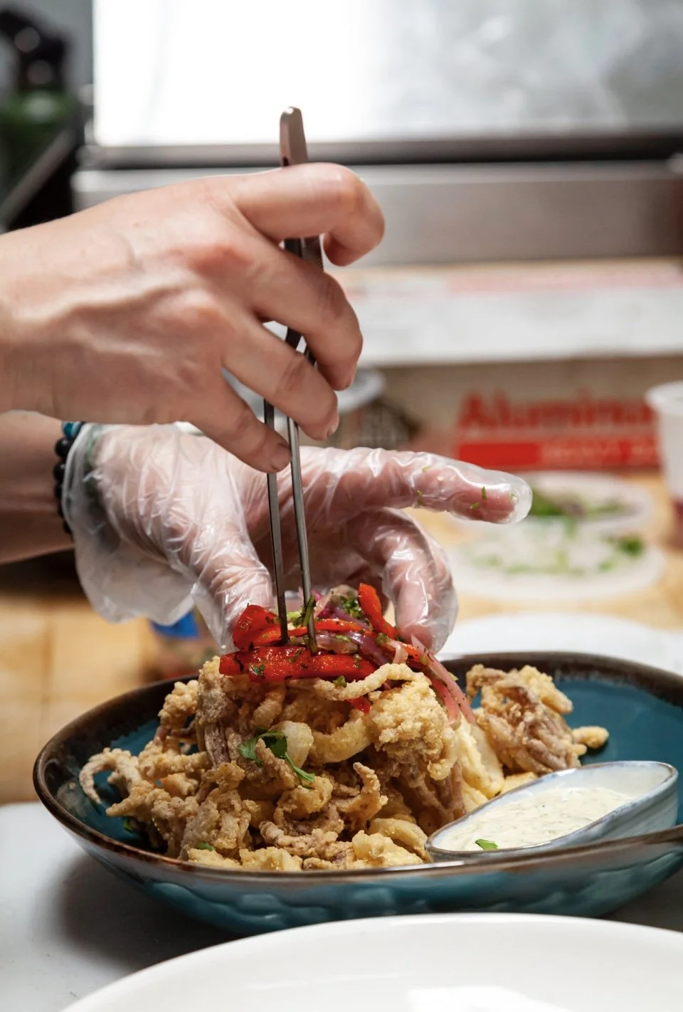 Close-up of a person's hand wearing a disposable glove and using tongs to add red chili peppers and cilantro on top of fried meat and noodles in a blue bowl, with a white sauce on the side.