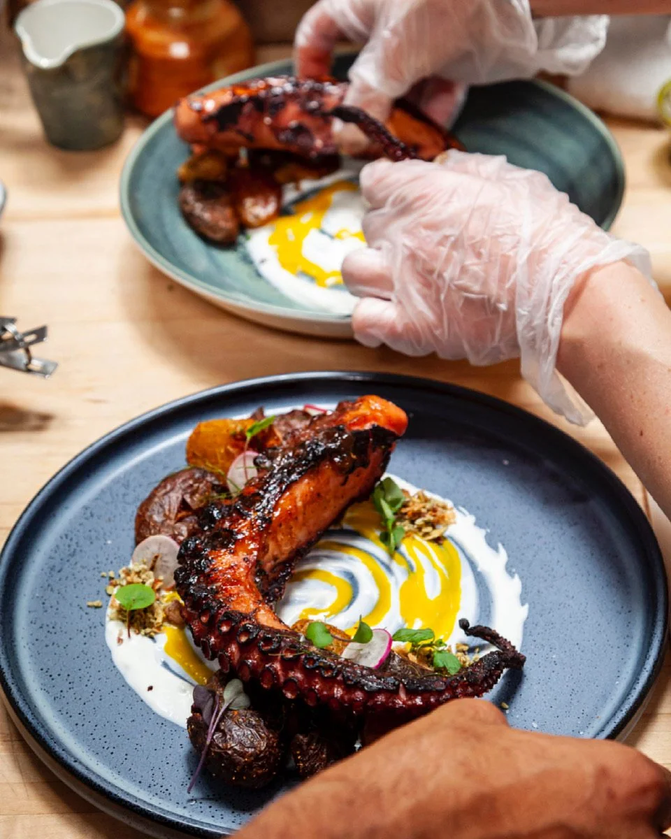 Chef preparing grilled octopus on a plate with sauce, garnishes, and vegetables, with another plate in the foreground and kitchen items in the background.