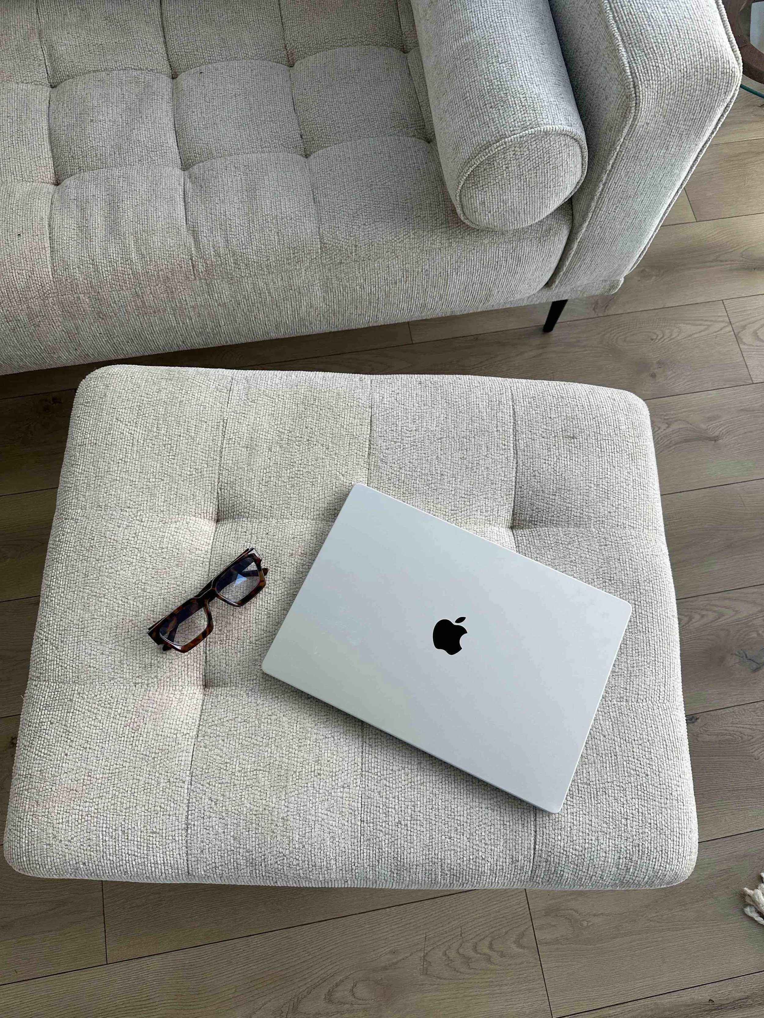 A beige upholstered ottoman with a closed silver MacBook and a pair of tortoiseshell glasses on top, with a gray sofa and wooden floor in the background.