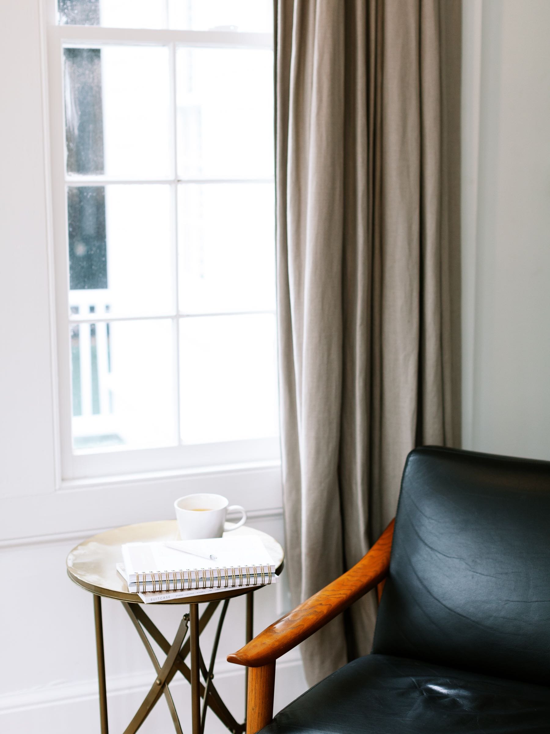 Black leather chair next to side table with journal, pen and coffee mug next to a big window with curtains