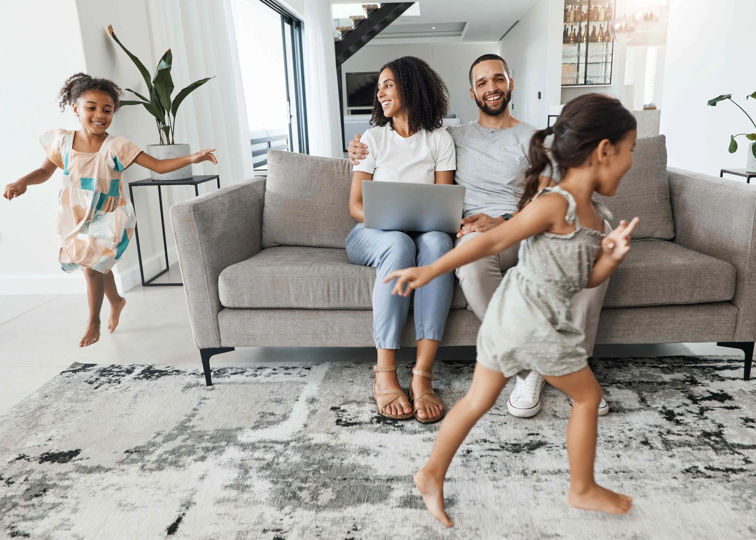 Young couple sitting on couch with laptop smiling while two children run around