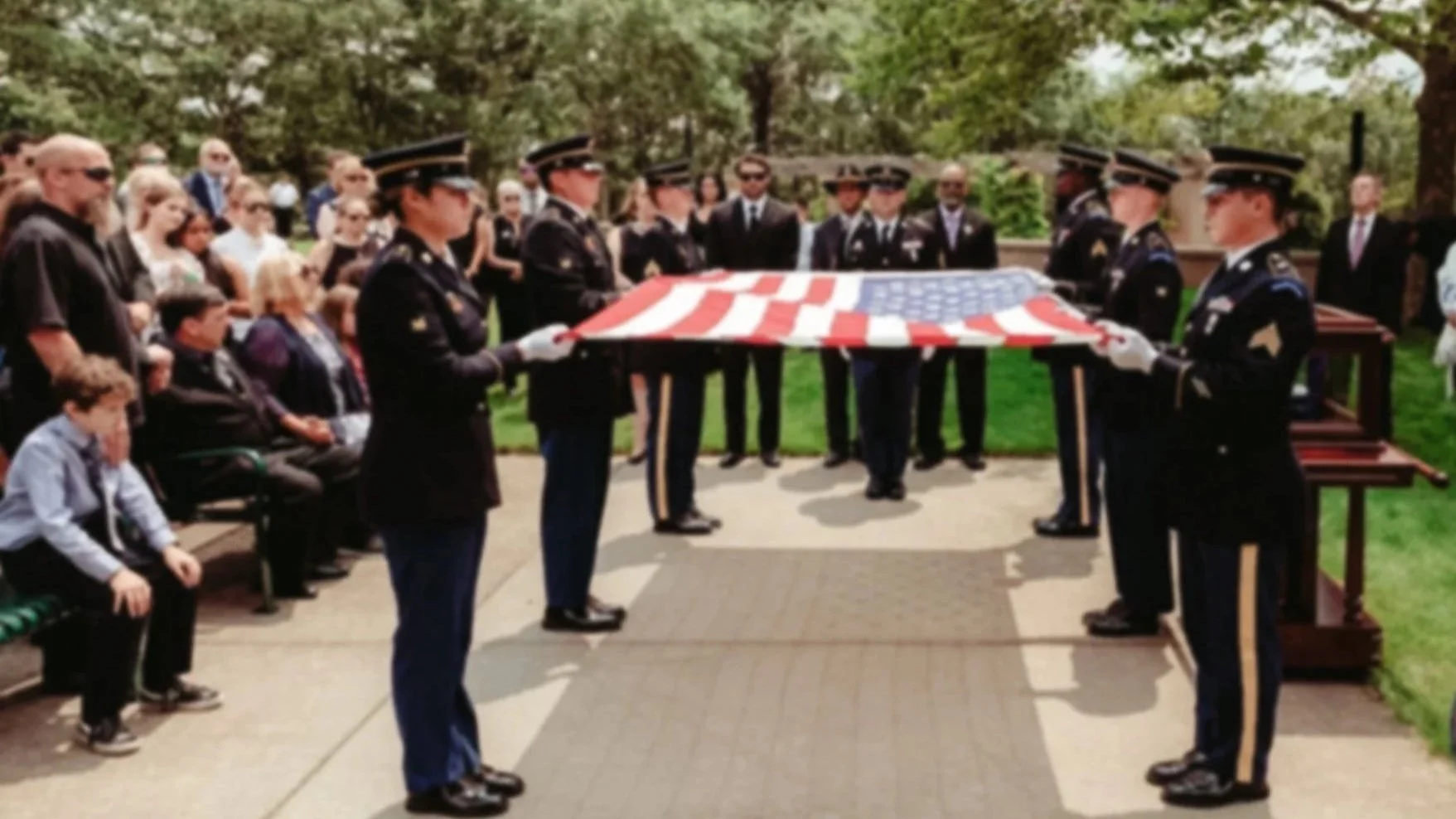 Military personnel in uniform fold an American flag during a solemn ceremony at a memorial site surrounded by onlookers and trees.