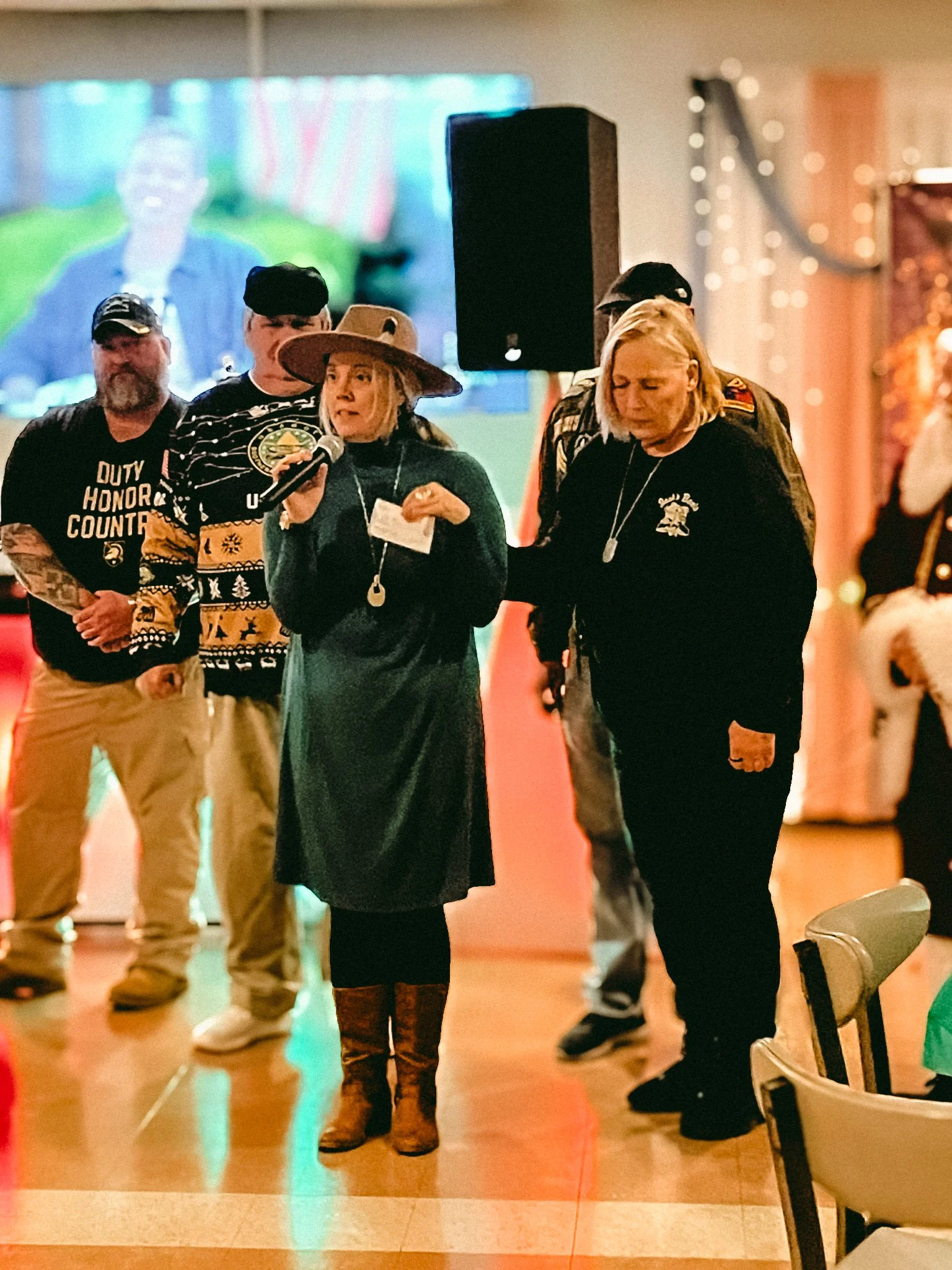 A group of people standing indoors at a social gathering. One woman is speaking into a microphone, wearing a tan hat and a dark dress with brown boots, while the others stand around her.