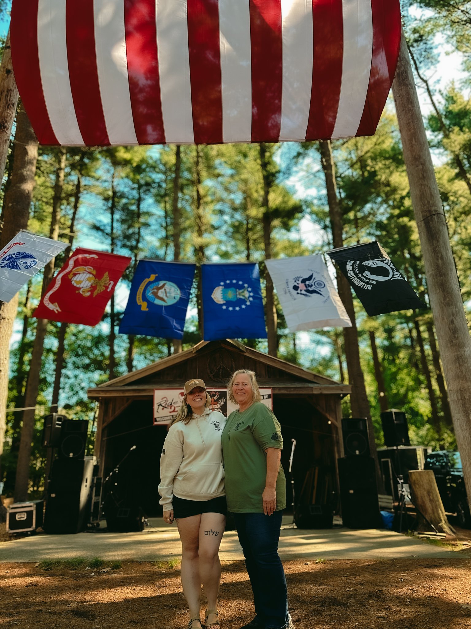 Two women standing close together outdoors in front of a stage with navy flags and a red, white, and blue striped canopy, surrounded by trees.