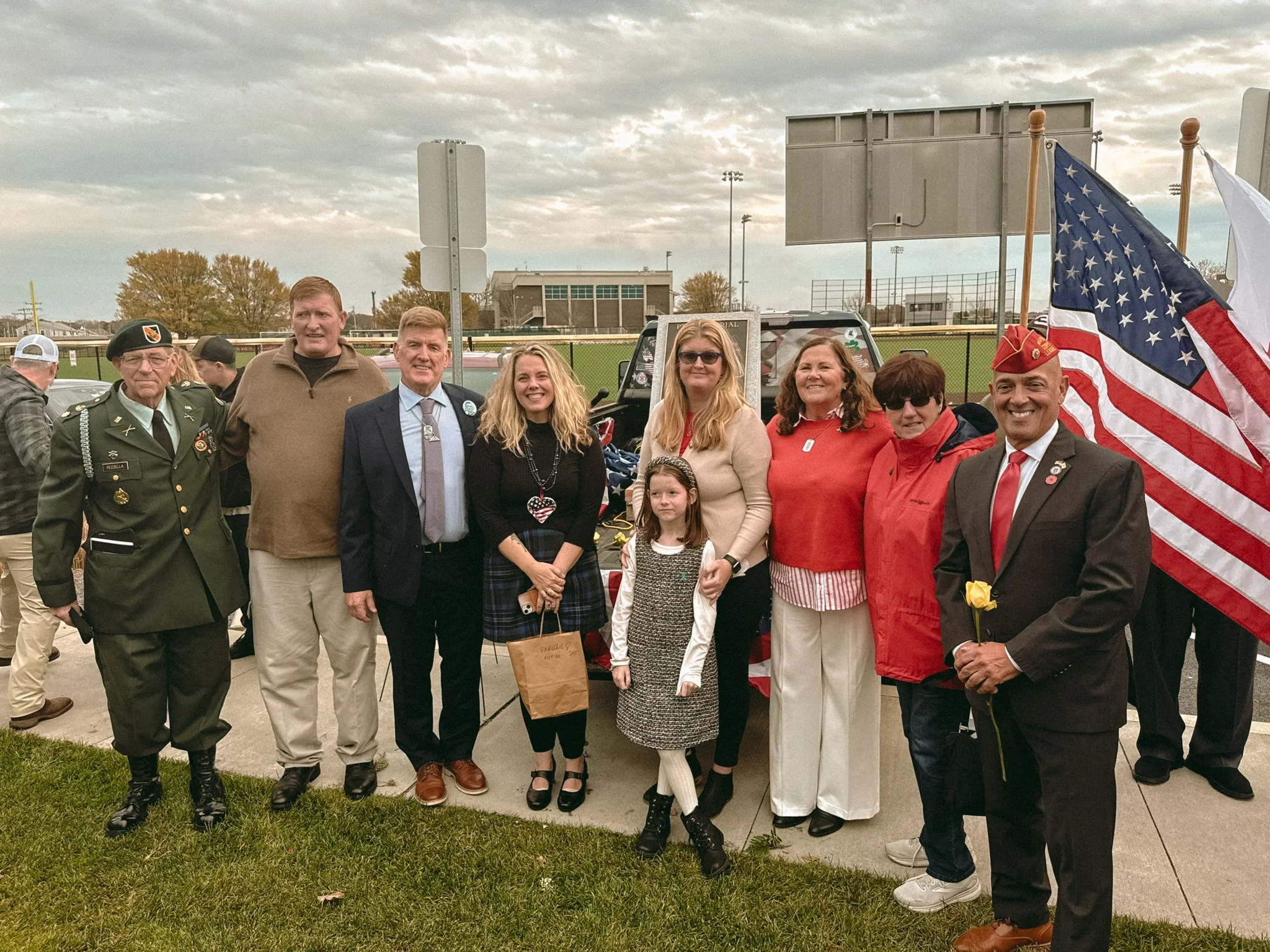 A group of diverse people, including veterans in uniform, civilians, and a young girl, standing outdoors on a cloudy day at a patriotic event. They are posing with American flags near a sports field.