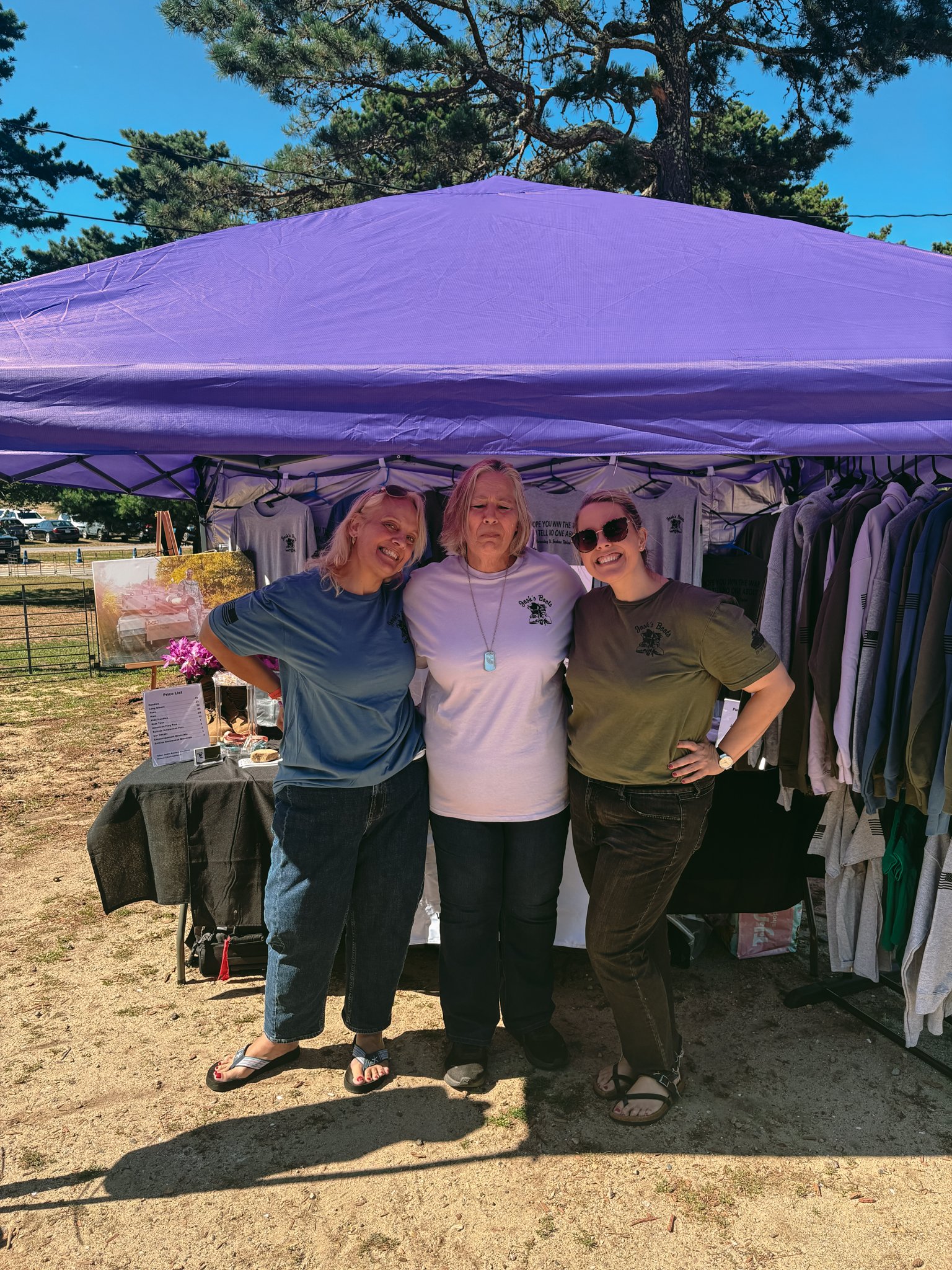Three women standing side by side under a purple canopy tent at an outdoor event. They are smiling and posing for the photo, with clothing and merchandise displayed behind them.