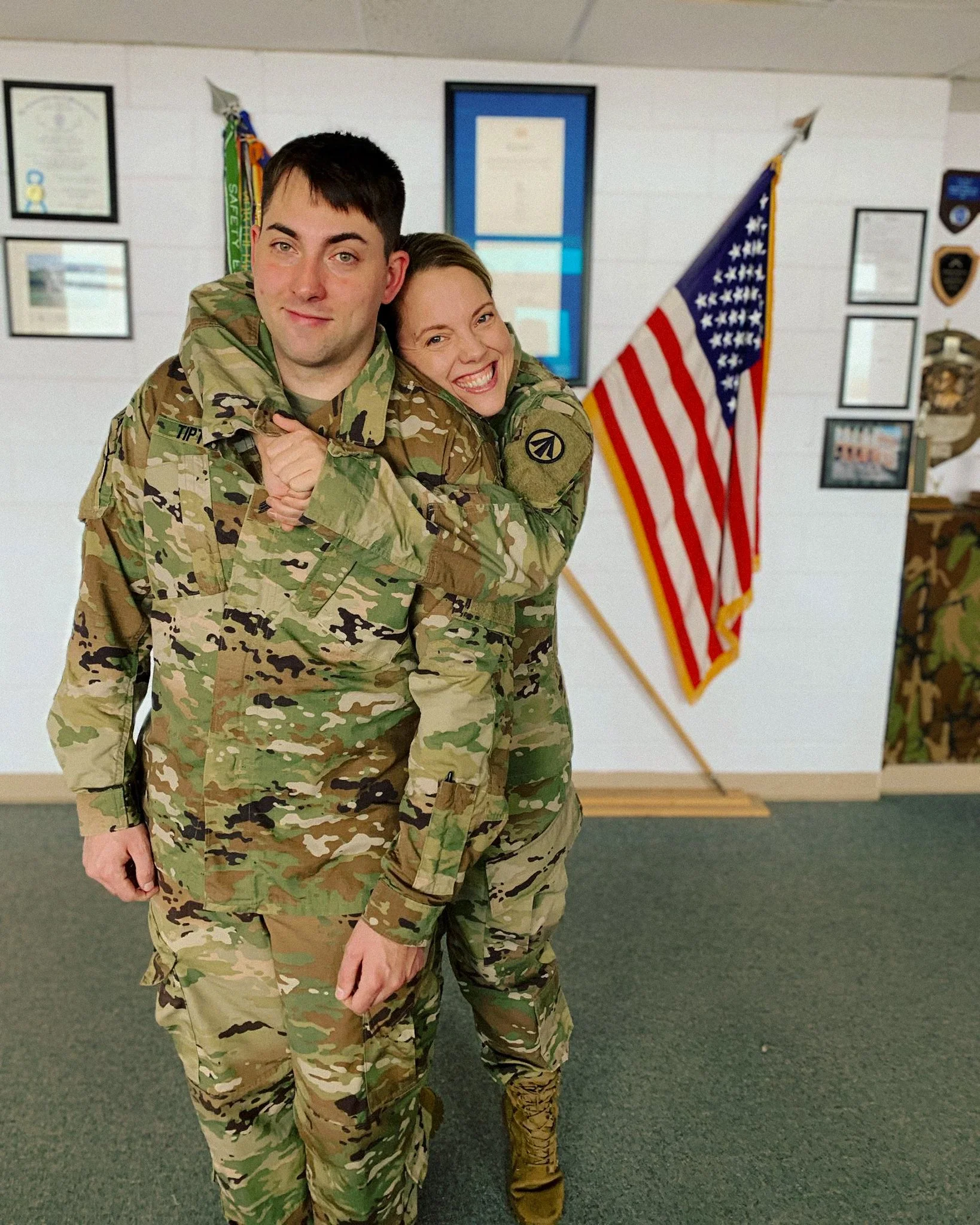 Siblings in camouflage military uniforms standing inside a room, hugging and smiling, with an American flag on a stand and framed certificates on the wall behind them.