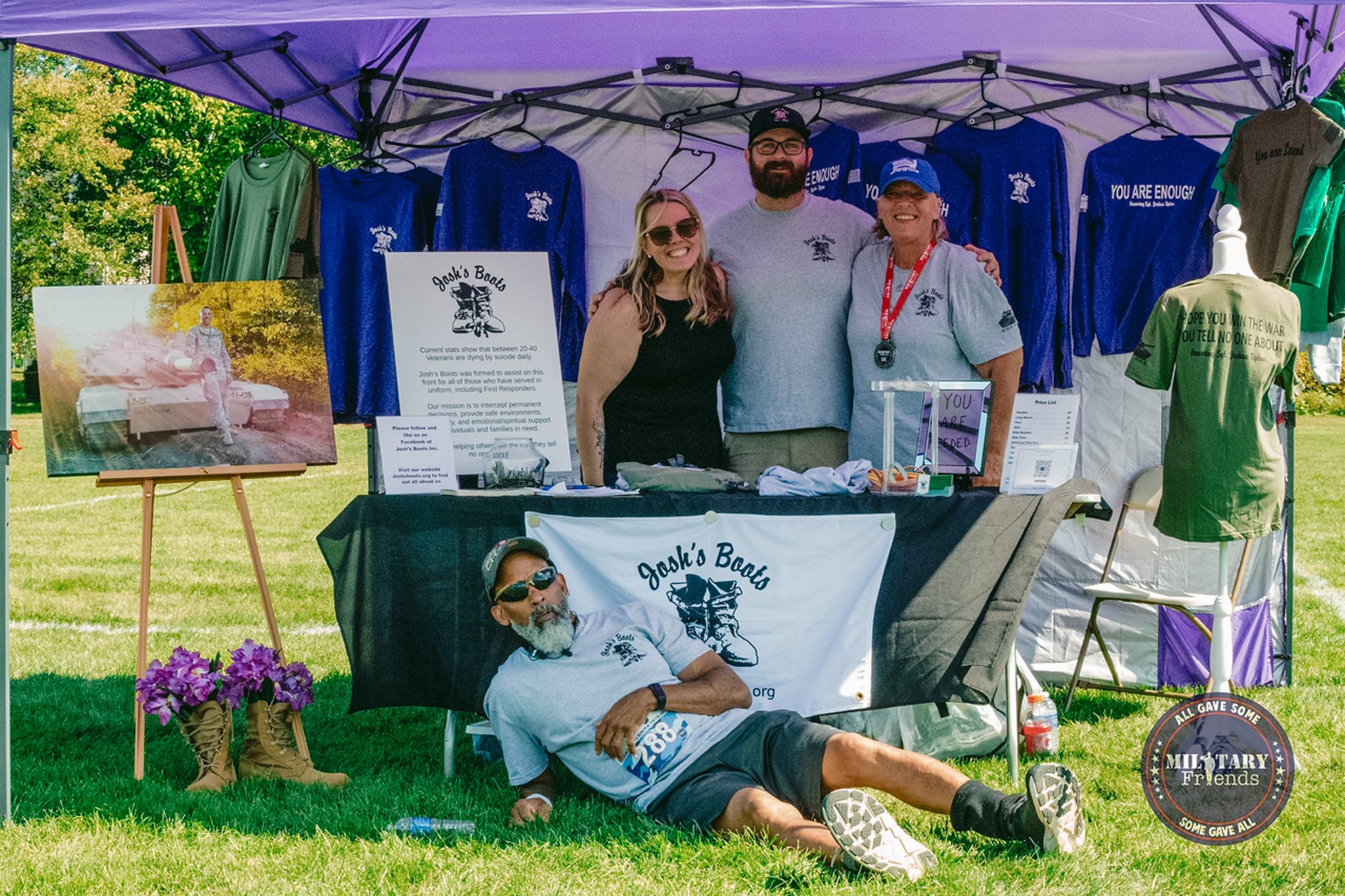 People standing at a booth for Joshi's Boots during an outdoor event, with T-shirts and posters displayed, and a man lying on the grass in front, wearing sunglasses and a shirt with the same logo.