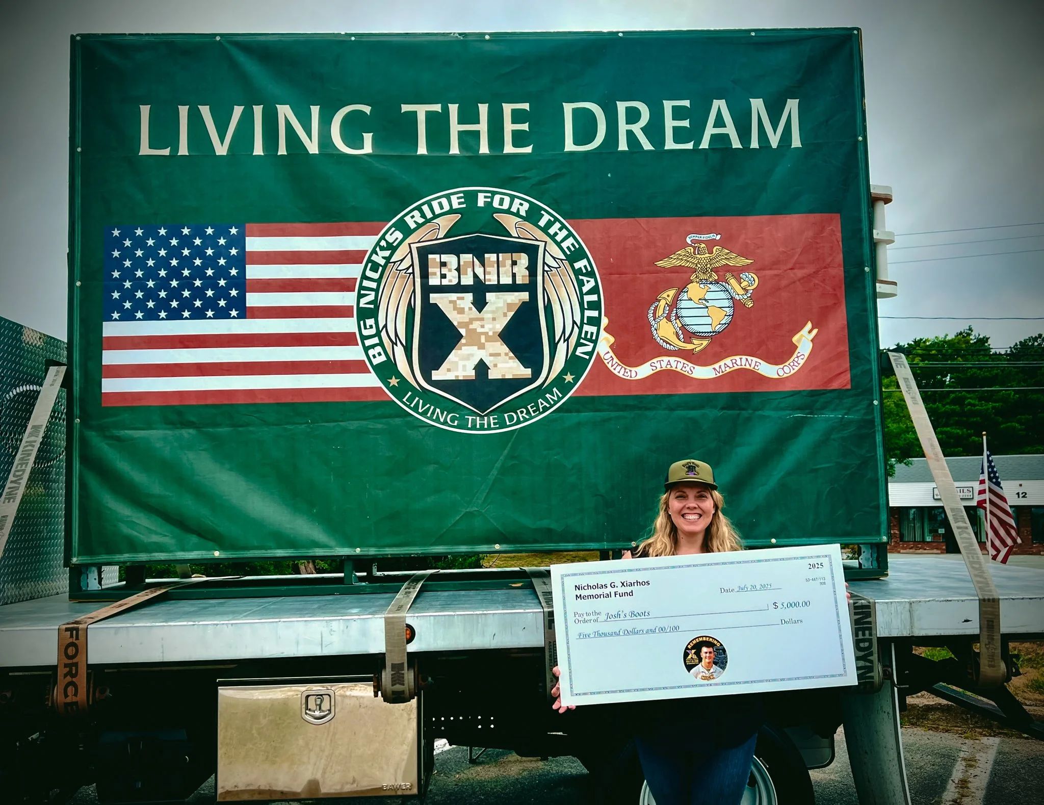 Woman in military hat holding large check in front of a military banner with the American flag and US Marine Corps emblem.