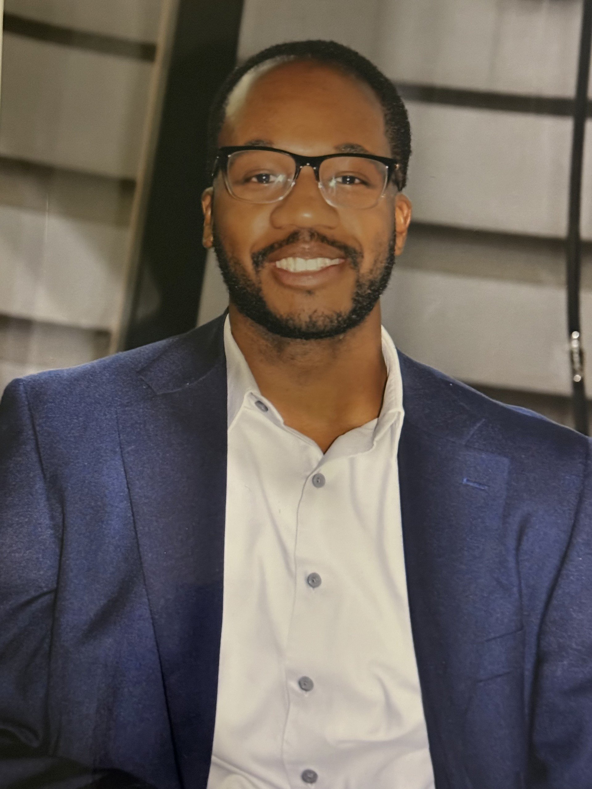 A man with glasses, a beard, and a mustache smiling, wearing a white dress shirt and a navy blazer.
