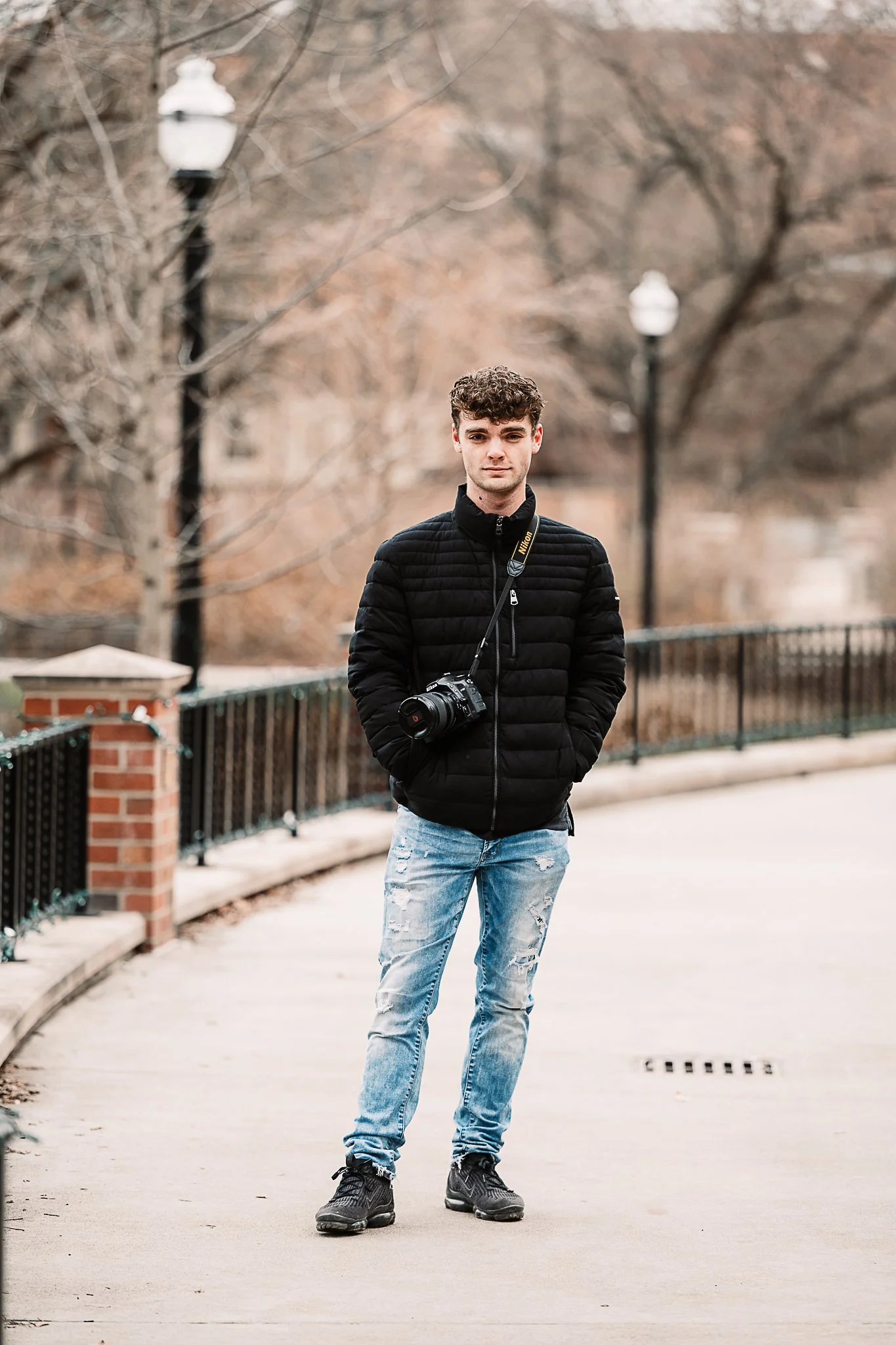 Young man in black jacket and ripped jeans standing on a sidewalk with his hands in his pockets, a camera hanging around his neck, and trees and street lamps in the background.