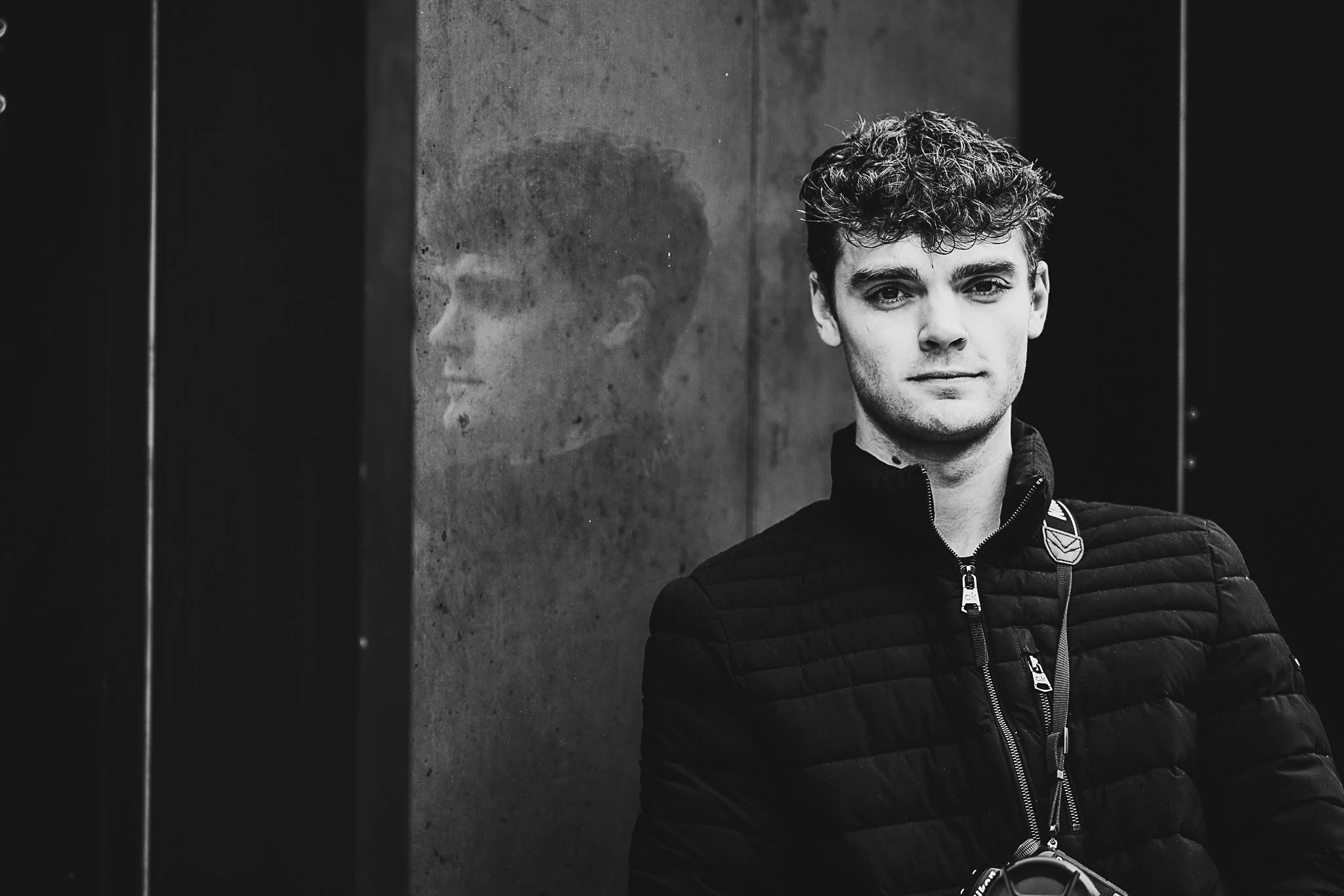 A young man with curly hair in a black quilted jacket standing against a concrete wall with his reflection visible in a mirror.