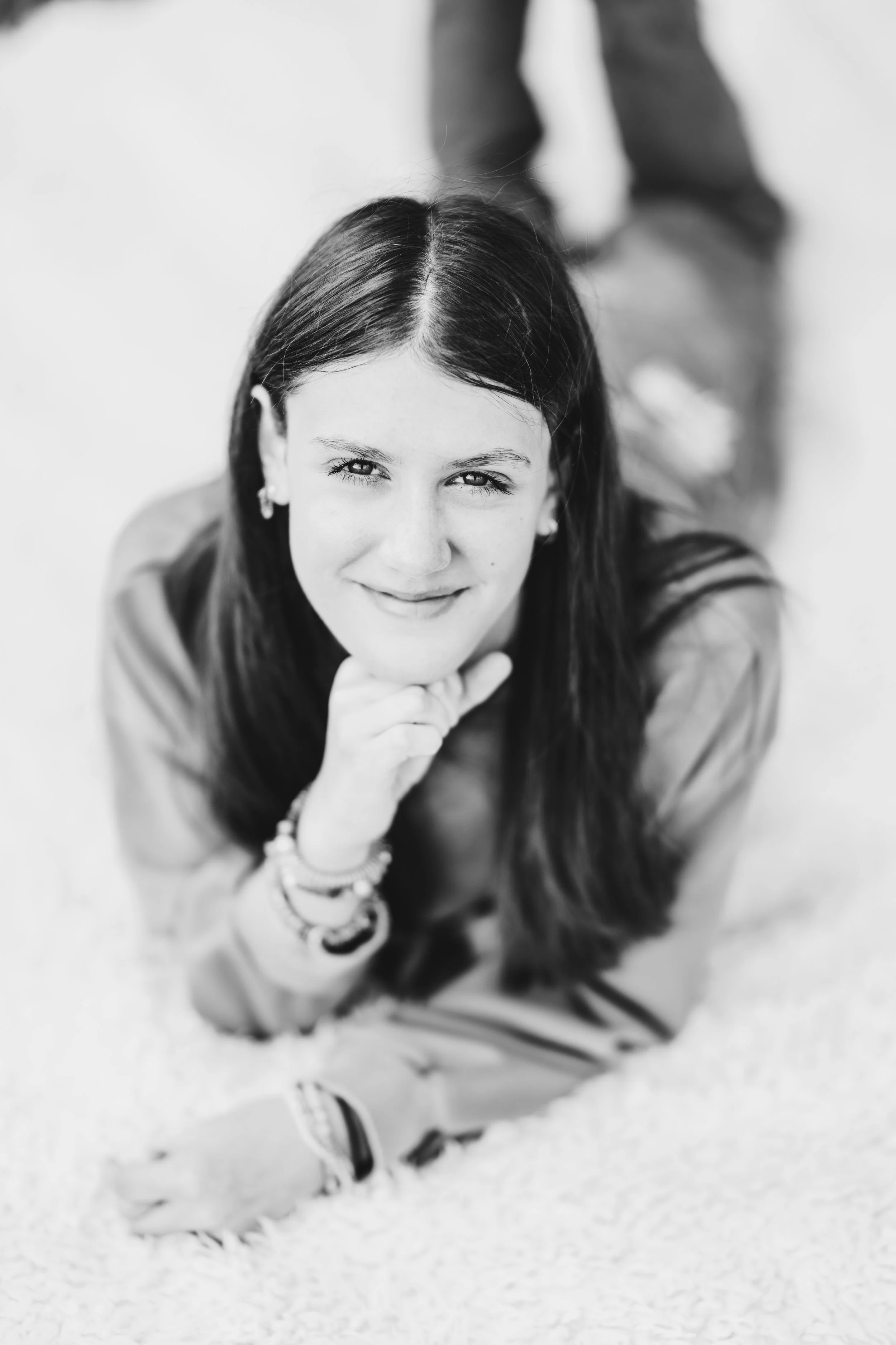 Black and white photo of a young woman lying on her stomach on a carpet, smiling, with her chin resting on her hand, and wearing bracelets and earrings.