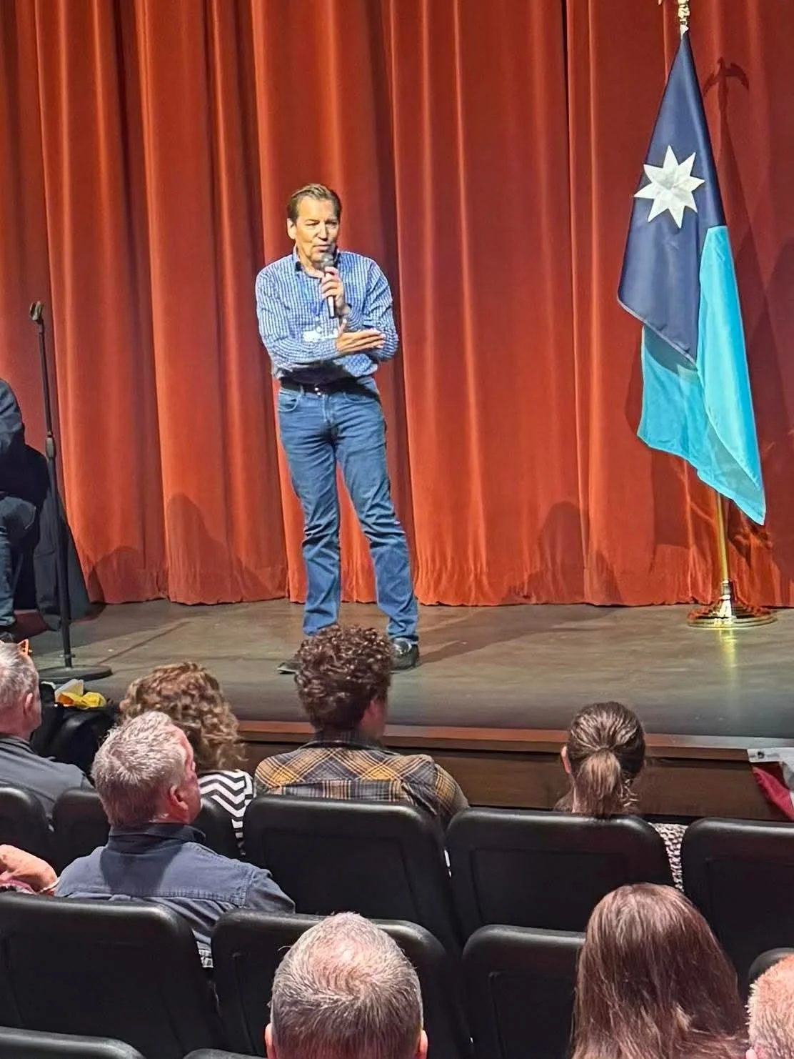 A man stands on a stage with red curtains, holding a microphone, in front of an audience. There is a flag standing next to him with a star emblem on a dark blue and light blue background.