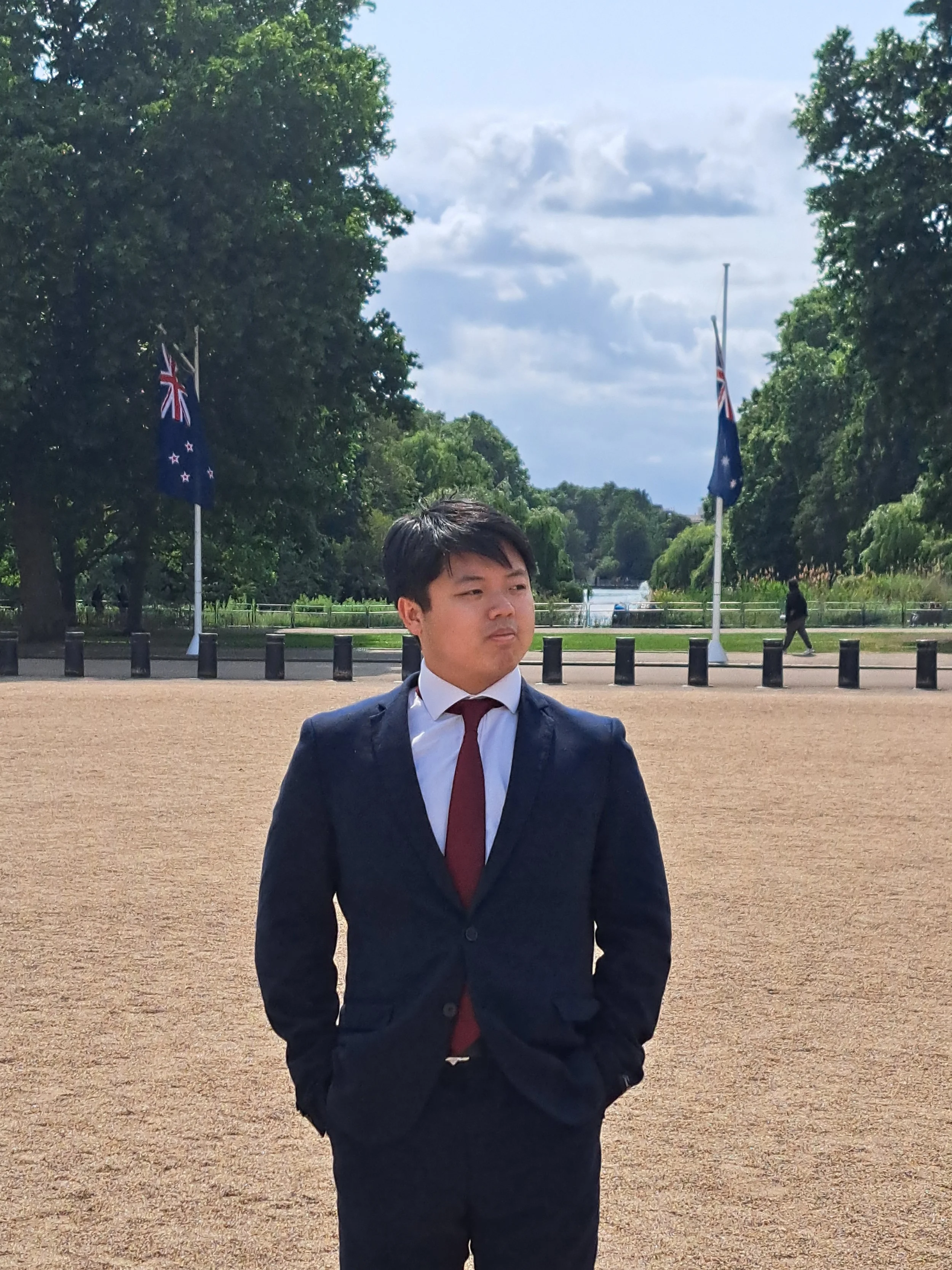 A young man in a navy suit, white shirt, and red tie stands outdoors on a sandy path, with greenery and trees behind him, and two flags on poles displayed in the background, under a partly cloudy sky.