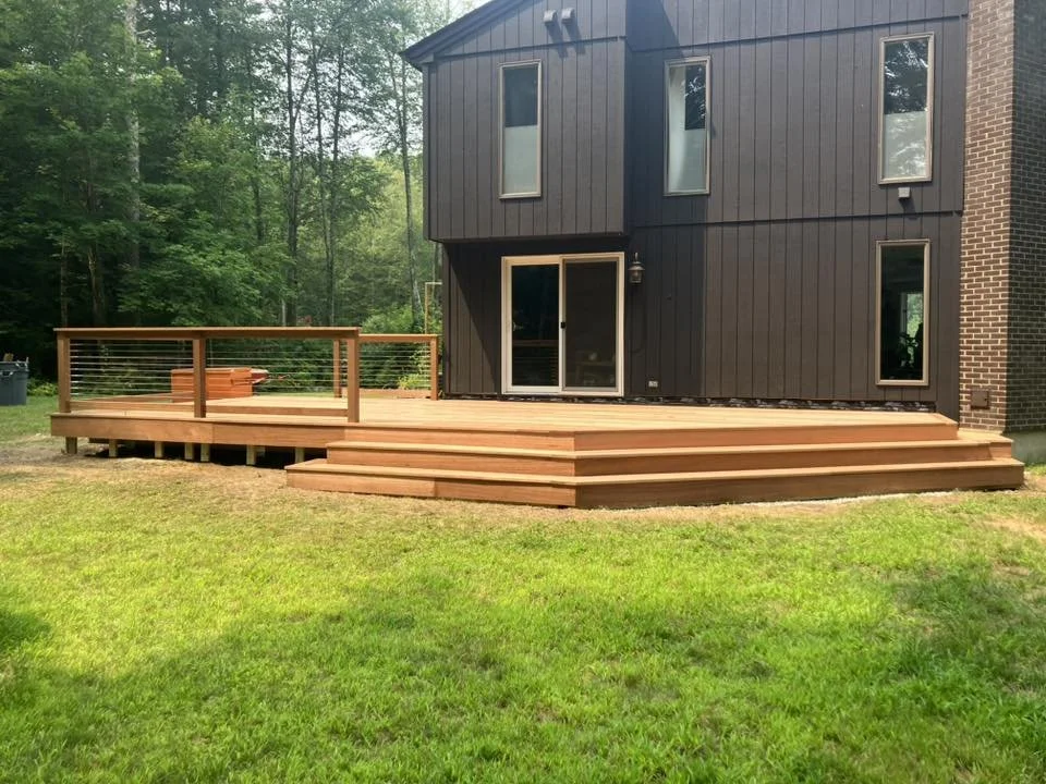 Newly built wooden deck with stairs and railing attached to a house, surrounded by a grassy lawn with trees in the background.