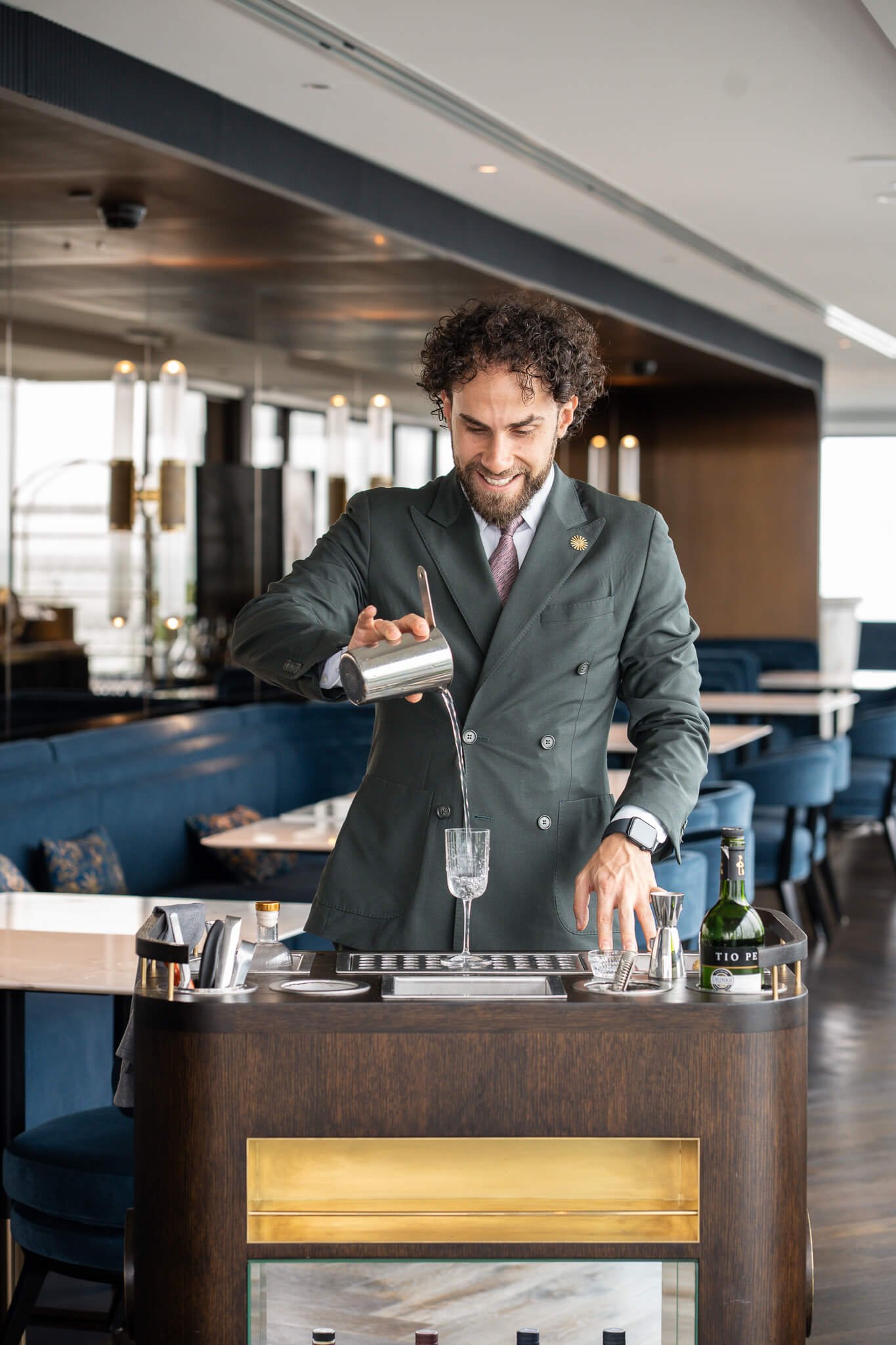 A bartender mixing a drink at a bar in a well-lit, modern restaurant. The bartender is smiling and wearing a dark suit and watch, pouring liquid from a metal shaker into a glass.