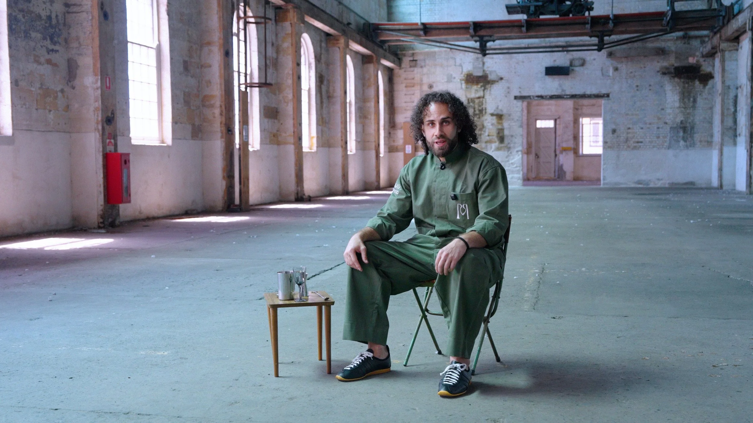 Man sitting on a chair in an empty, industrial-style room with large windows, wearing a green jumpsuit, with a small wooden table holding a cocktails mixing tin in front of him.