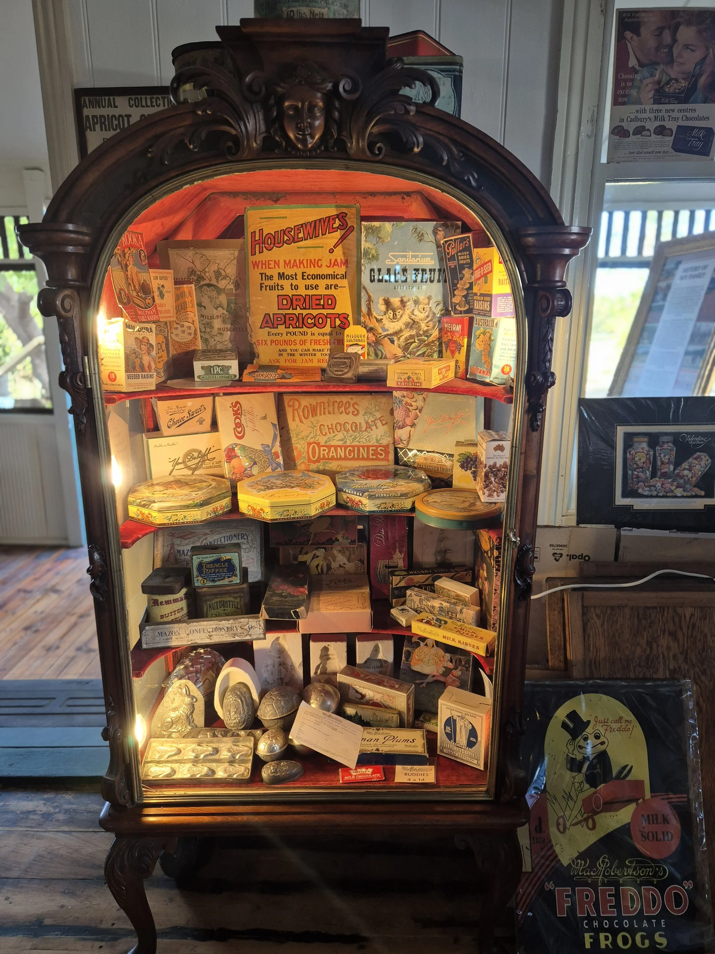 An ornate wooden display cabinet filled with vintage candy and confectionery items, including boxes of chocolates, tins, and novelty sweets, with a sign about dried apricots and other old-fashioned treats inside.