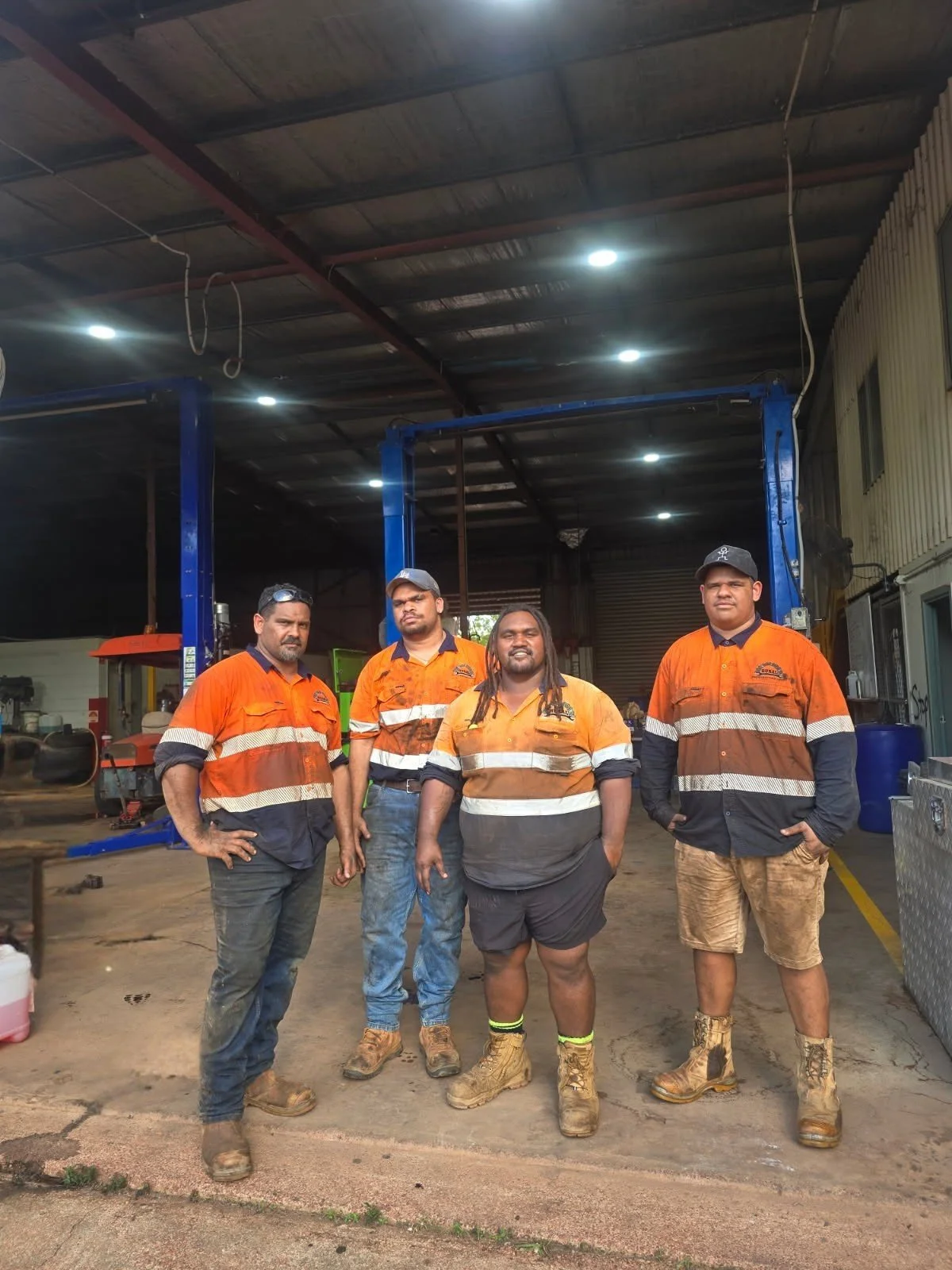 Four workers standing together inside a workshop, all wearing high-vis orange safety uniforms and work boots.