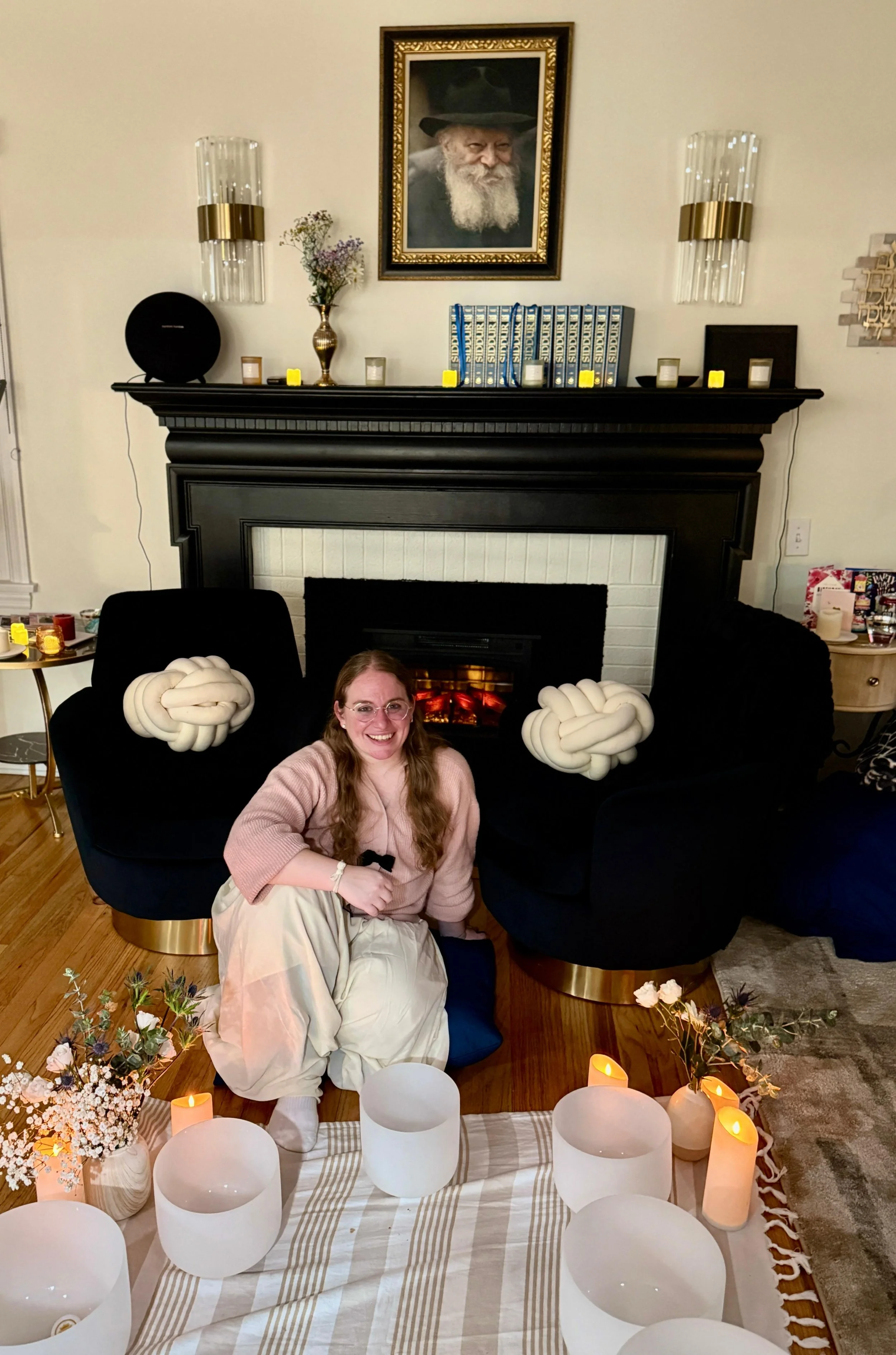 A woman sitting cross-legged on the floor in front of a fireplace, smiling, with decorative pillows resembling brain shapes on black chairs on either side. The room has a decorated mantel with candles, a framed photo of an elderly man, and various decorative items, with candles arranged on a striped cloth in front of the woman.