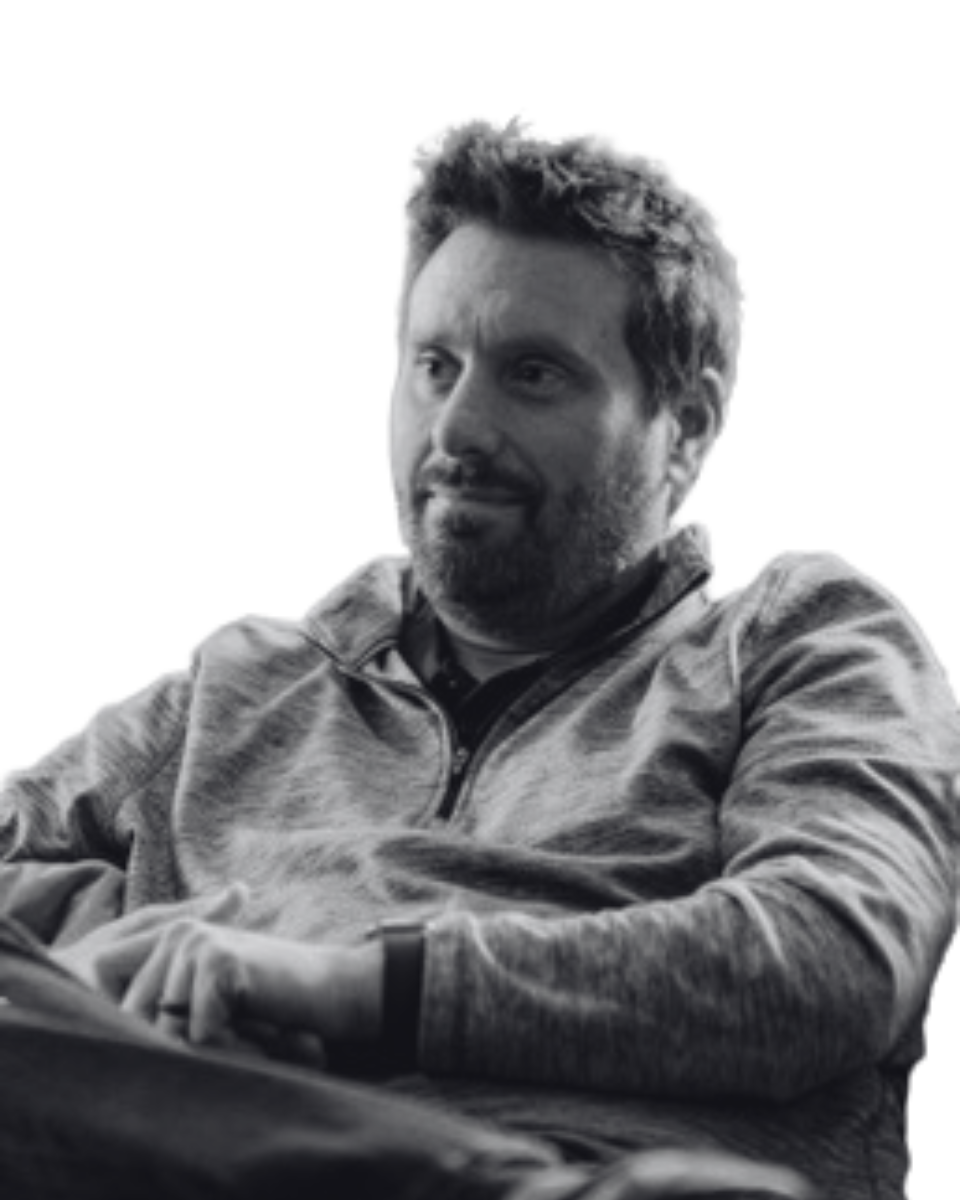 Black and white photo of a man with a beard and short hair, sitting and smiling slightly, wearing a casual zip-up jacket.