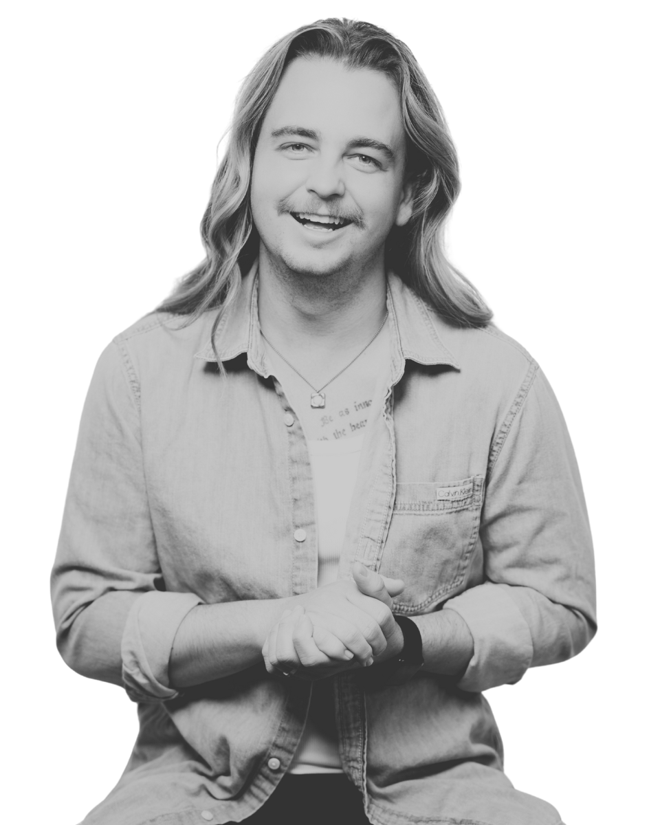 Black and white photo of a smiling young man with long hair, wearing a denim shirt and a necklace, sitting against a plain background.