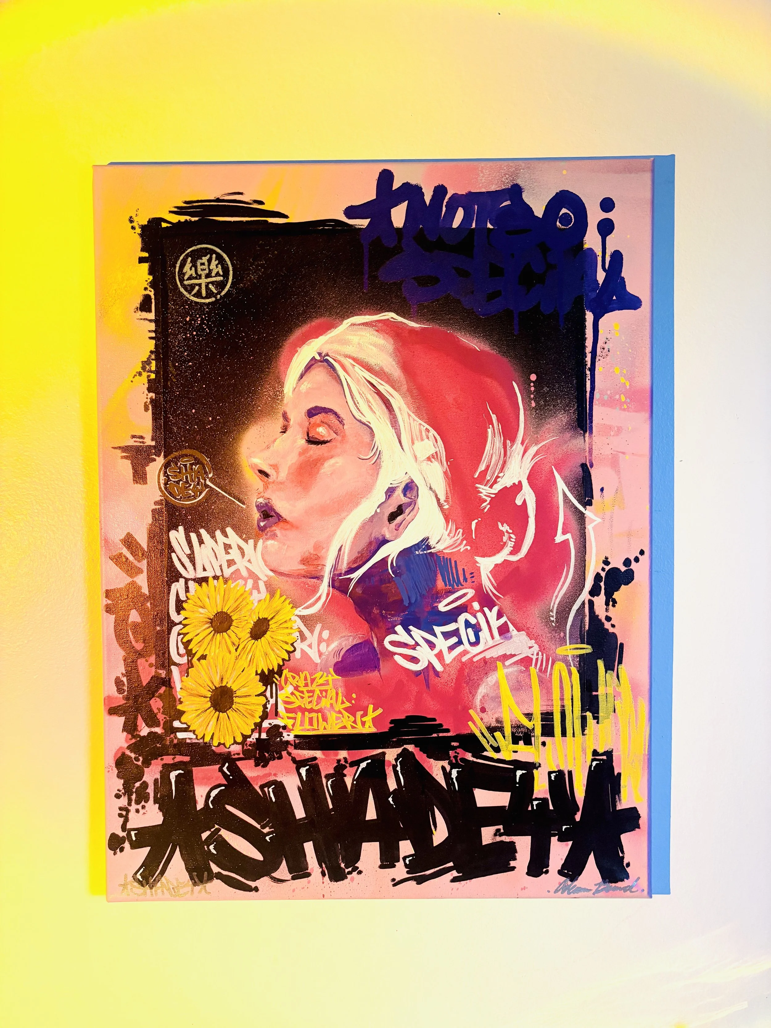 Street art mural of a woman's face with closed eyes, pink hair, surrounded by graffiti tags, yellow flowers, and colorful background.