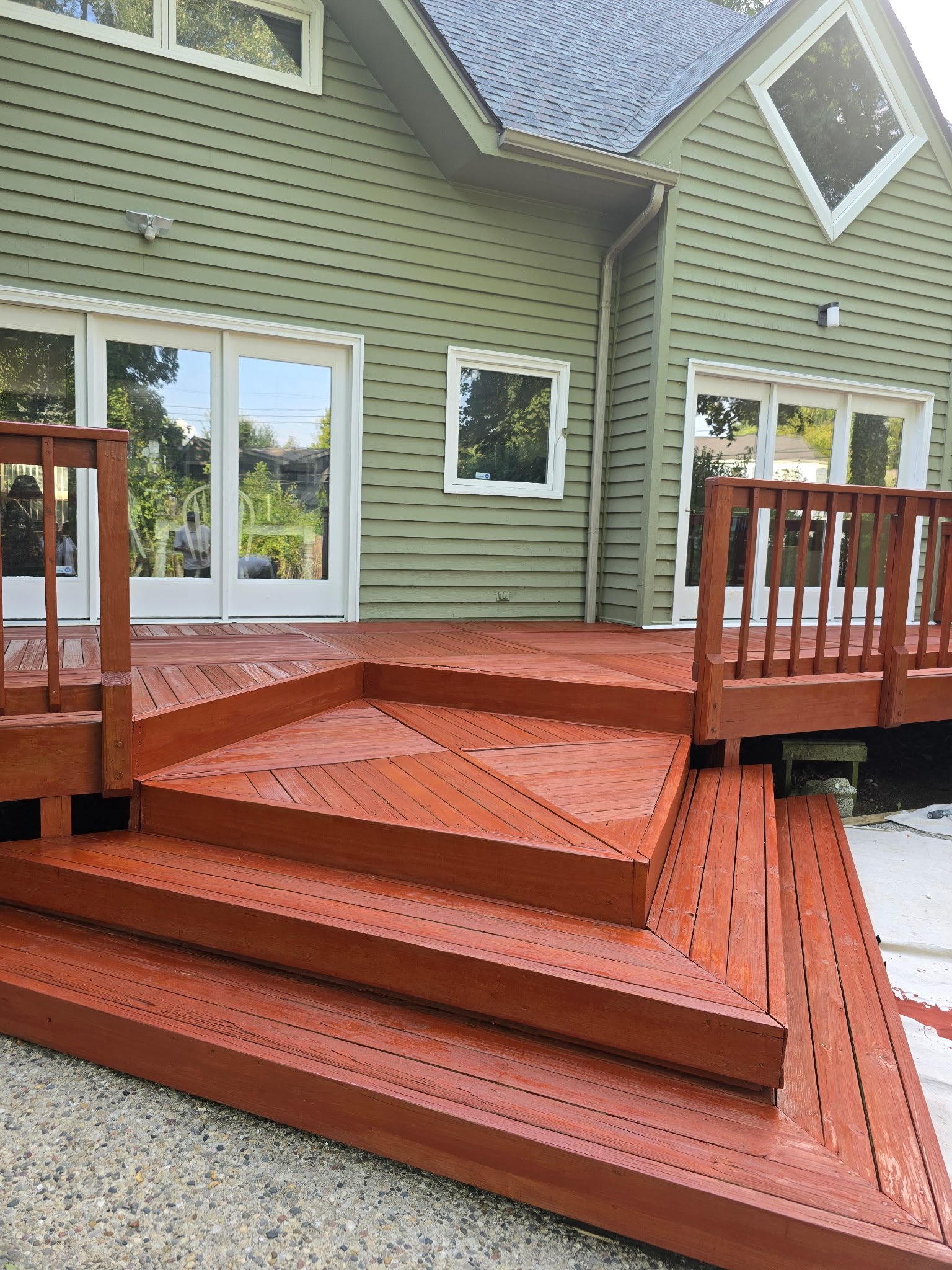 Newly built red wooden deck attached to a green house with large windows and sliding glass doors; the deck has multiple levels and a geometric pattern, with a railing on one side.