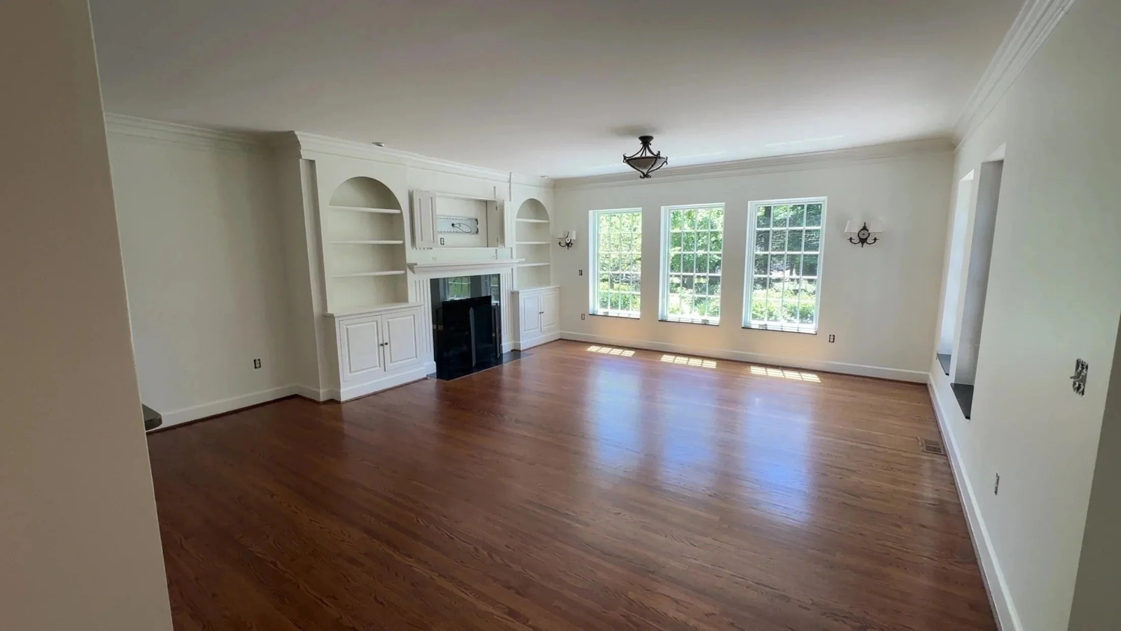 Empty living room with hardwood floors, white walls, built-in shelves and cabinet around a fireplace, three large windows with natural light, and ceiling light fixtures.