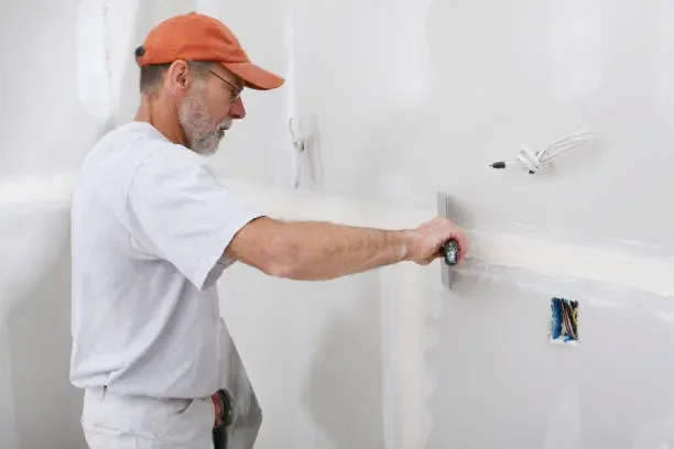 A man wearing an orange cap, glasses, and a white t-shirt is smoothing drywall with a trowel while working on a wall with exposed electrical wiring.