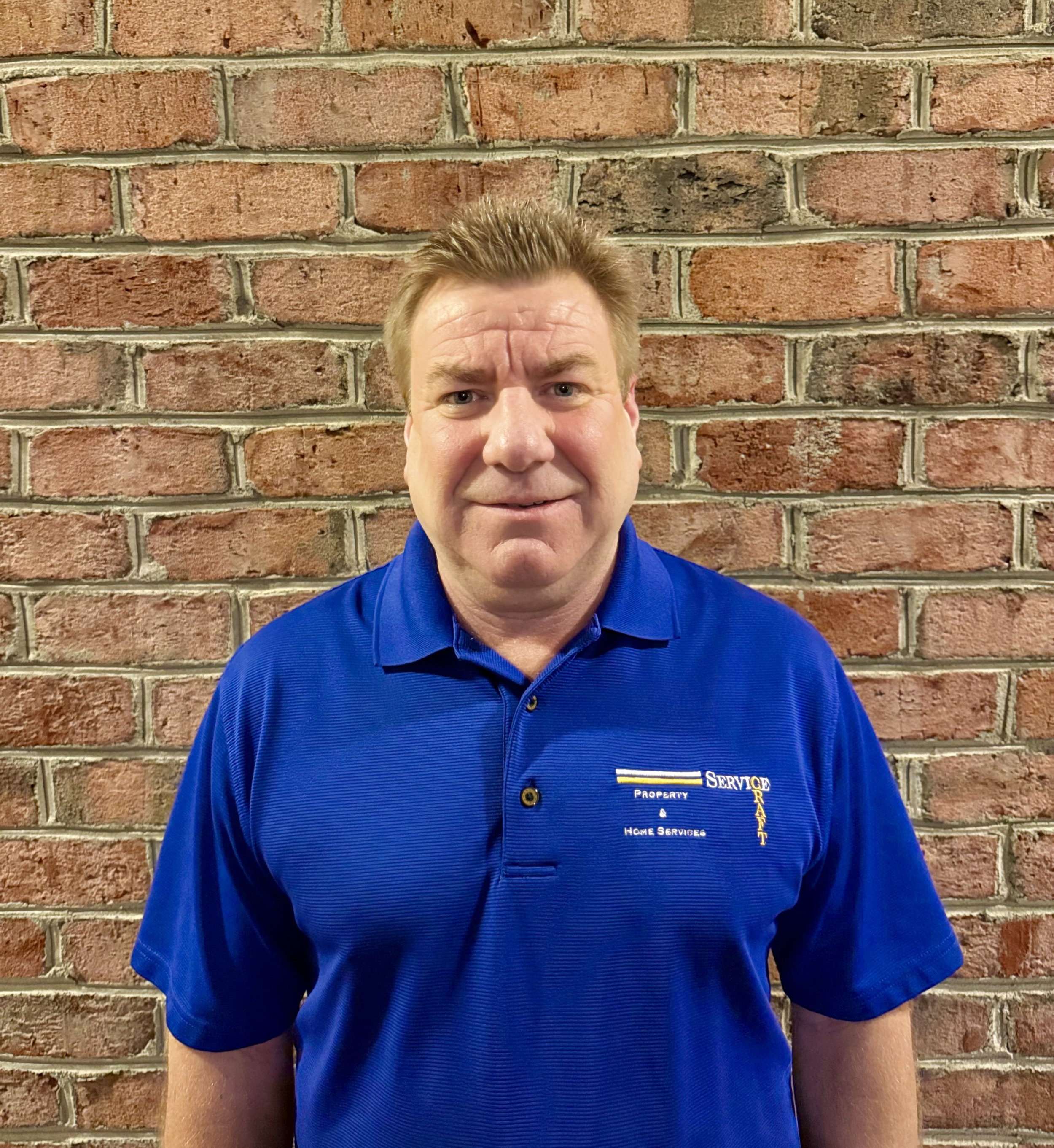 A man with light skin, short blonde hair, wearing a blue polo shirt with a logo that reads 'Property & Home Services' and 'Service Ready', standing in front of a red brick wall.