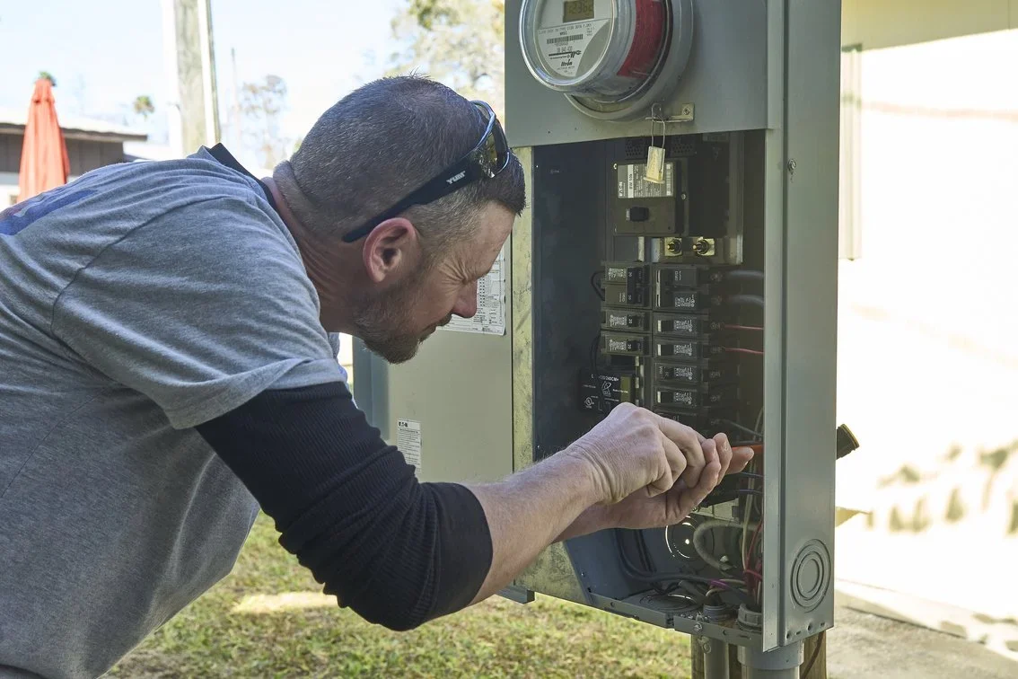 A man working on an outdoor electrical meter box, inspecting and fixing the circuit breakers.