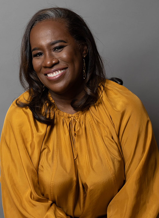 Portrait of a smiling African American woman with dark hair in loose waves, wearing a mustard yellow blouse with a gathered neckline and gold hoop earrings, against a plain gray background.
