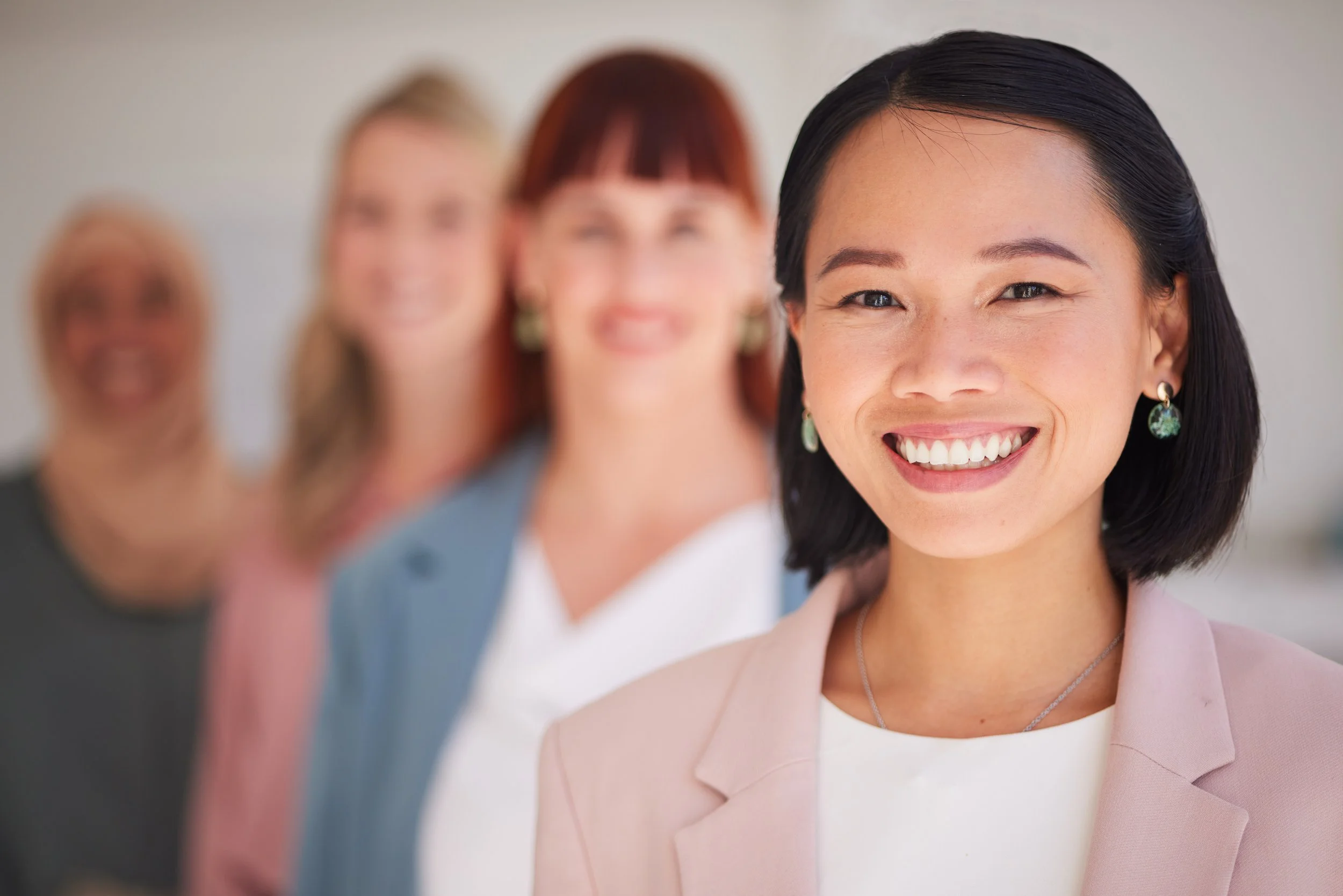 Four women standing in a row, smiling in a bright room. The woman in the front is Asian, wearing a light pink blazer and earrings. The other women are blurred in the background.
