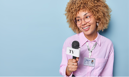 A woman with curly hair, glasses, and a name badge, holding a microphone labeled 'TV' and smiling against a blue background.