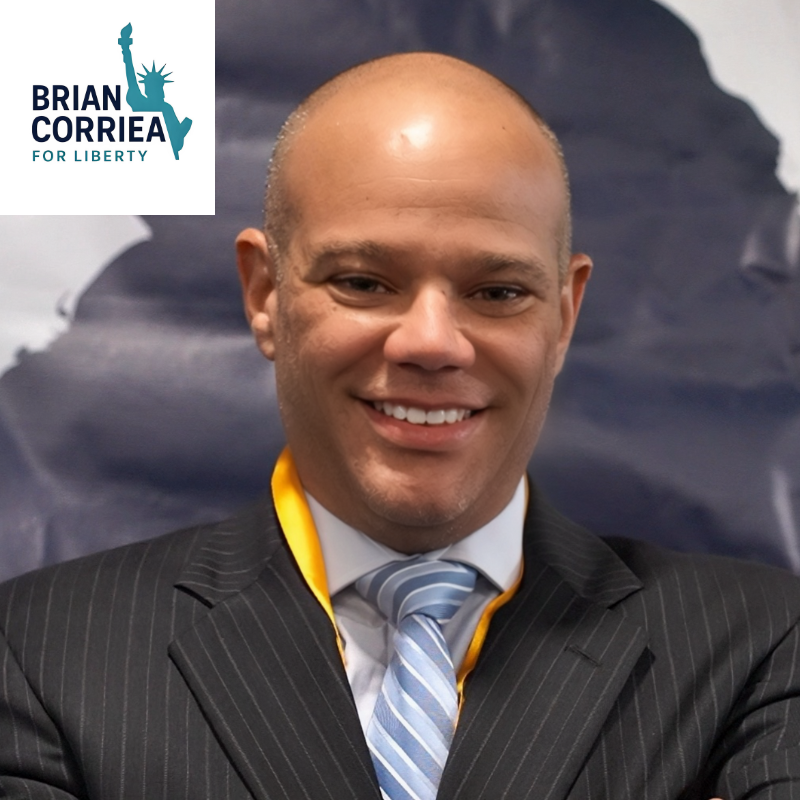 Photograph of a smiling man in a suit and tie, with a lapel medal, standing in front of a background with the logo 'Brian Corriea for Liberty' featuring an outline of the Statue of Liberty.