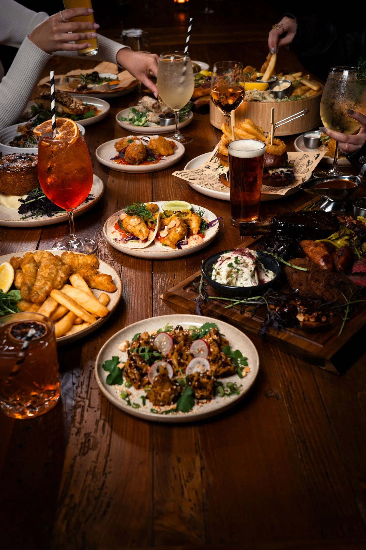 A variety of appetizer and main dishes on a wooden table, including fried items, salads, and grilled meats, with people reaching for food and drinks.