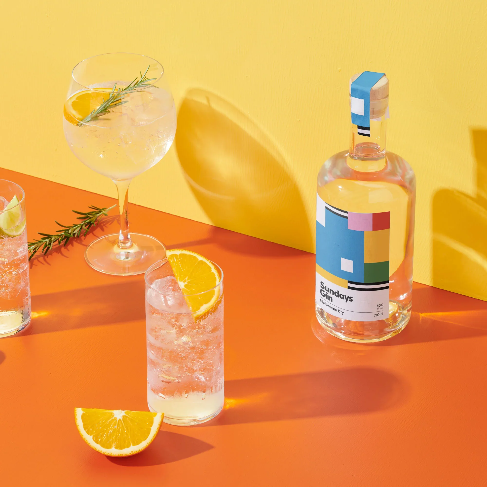 A tall glass of gin and tonic garnished with lemon slices and fresh rosemary, next to a half lemon. In the background, a bottle of Sunday Gays gin with a colorful label and wooden cap, against a yellow wall and orange table.