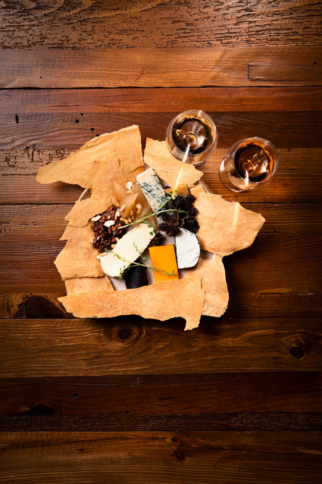 Cheese platter with various cheeses, nuts, bread crisps, and black grapes, served with two glasses of rosé wine on a wooden table.